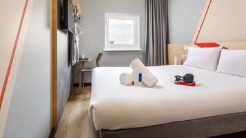 A hotel room with a bed, a desk, a chair, a window, and a TV mounted on the wall.