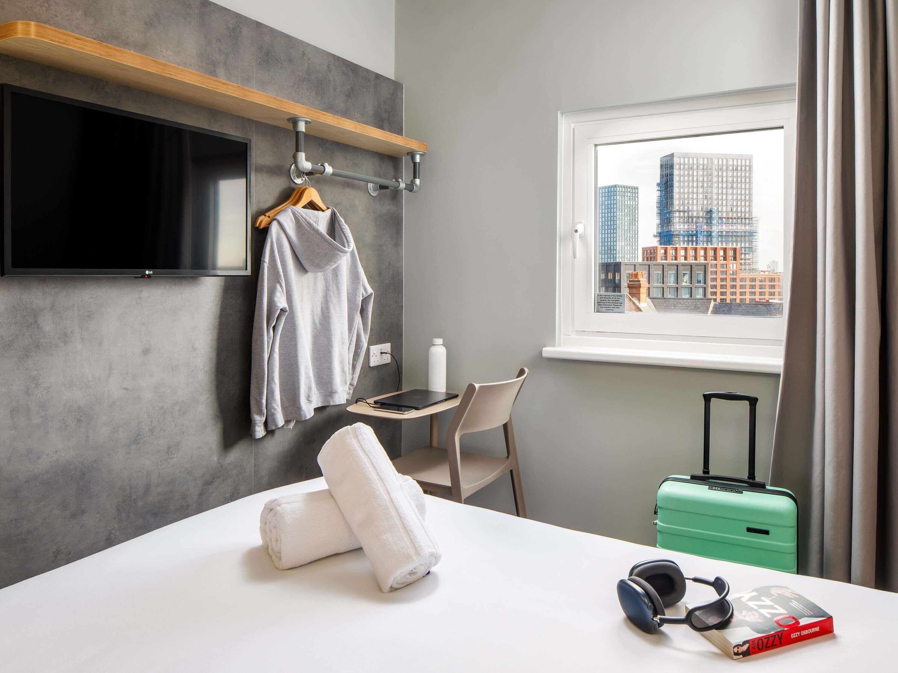 A hotel room with a bed, a TV, a window with a city view, a desk with a chair, a suitcase, and a book.