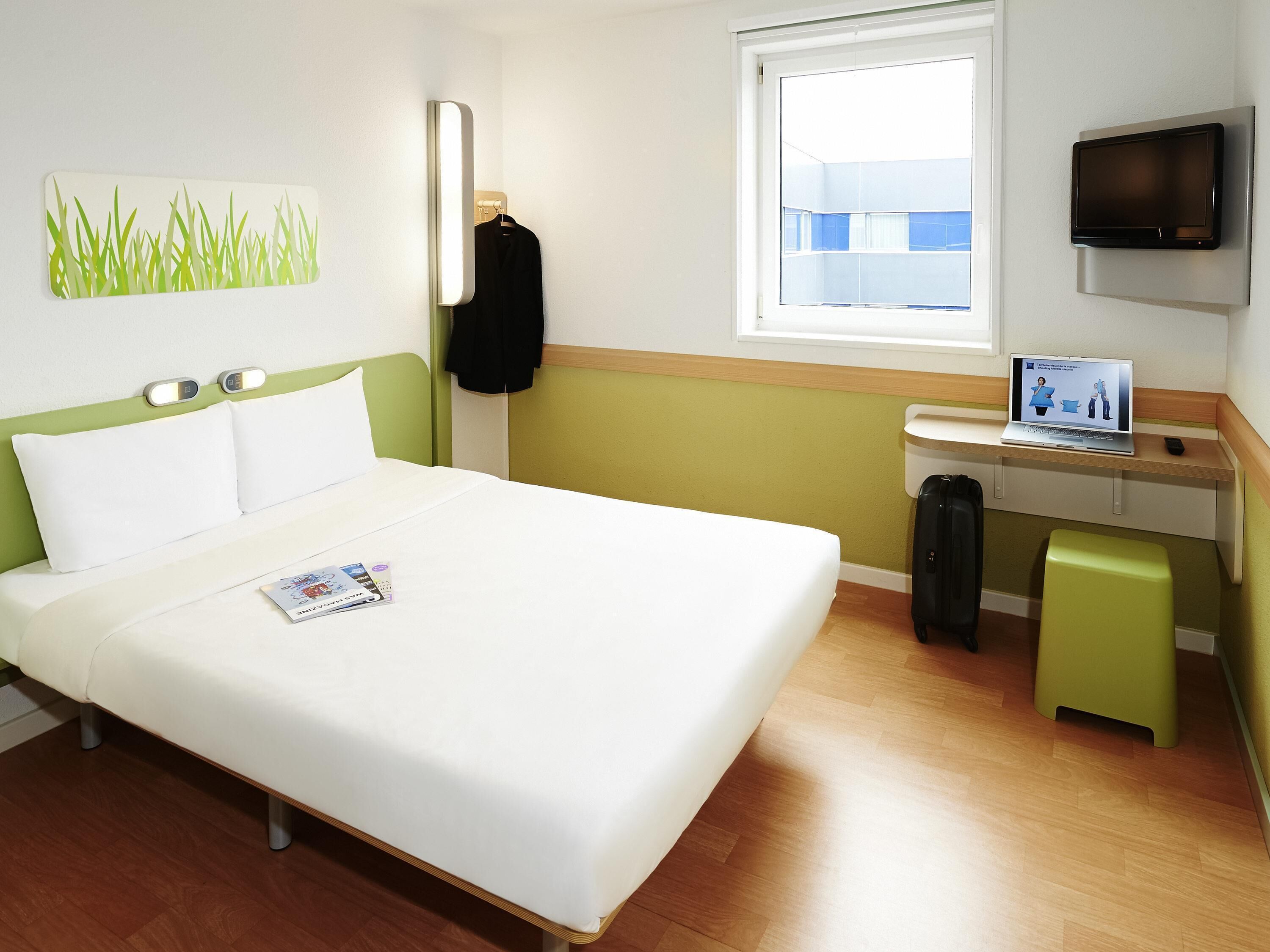 A hotel room with a bed, a desk with a laptop, a TV, and a window.