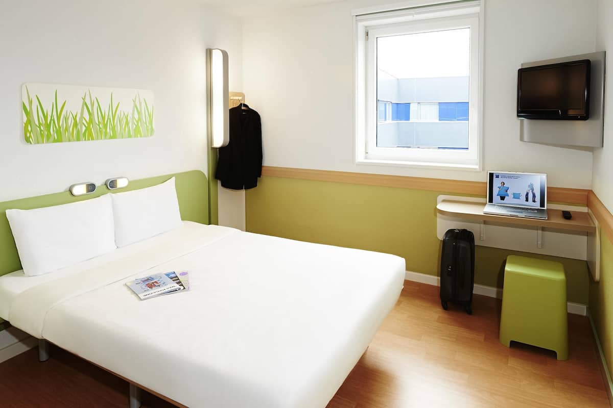 A hotel room with a bed, a desk with a laptop, a TV, and a window.