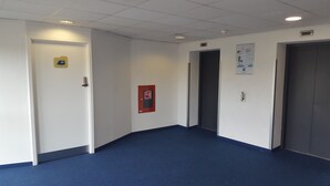 Un pasillo con alfombra azul, paredes blancas y puertas que dan a habitaciones. Hay un detector de incendios en la pared y un cartel a la derecha.
