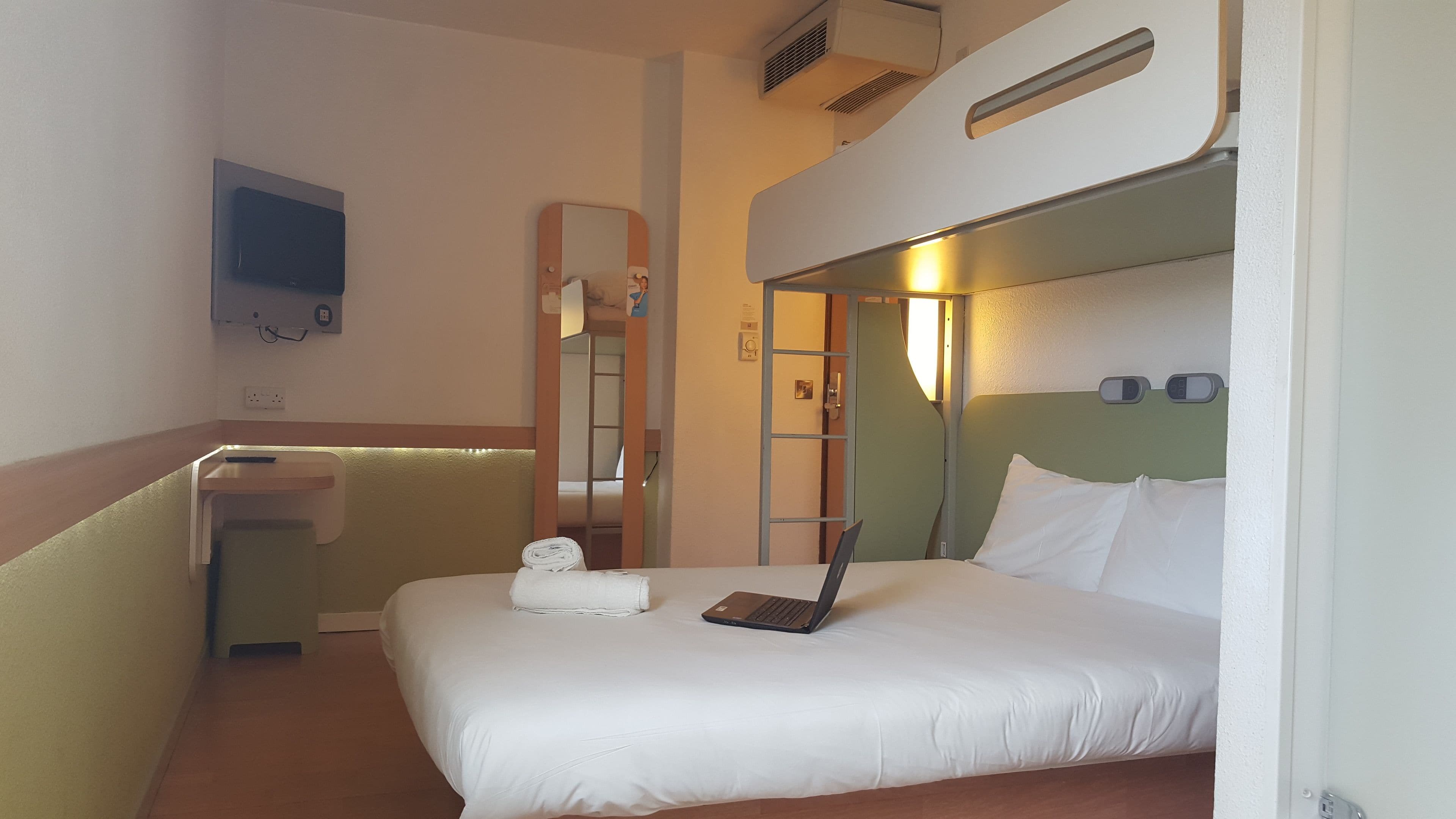 A hotel room with a bed, a desk, a mirror, and a laptop.