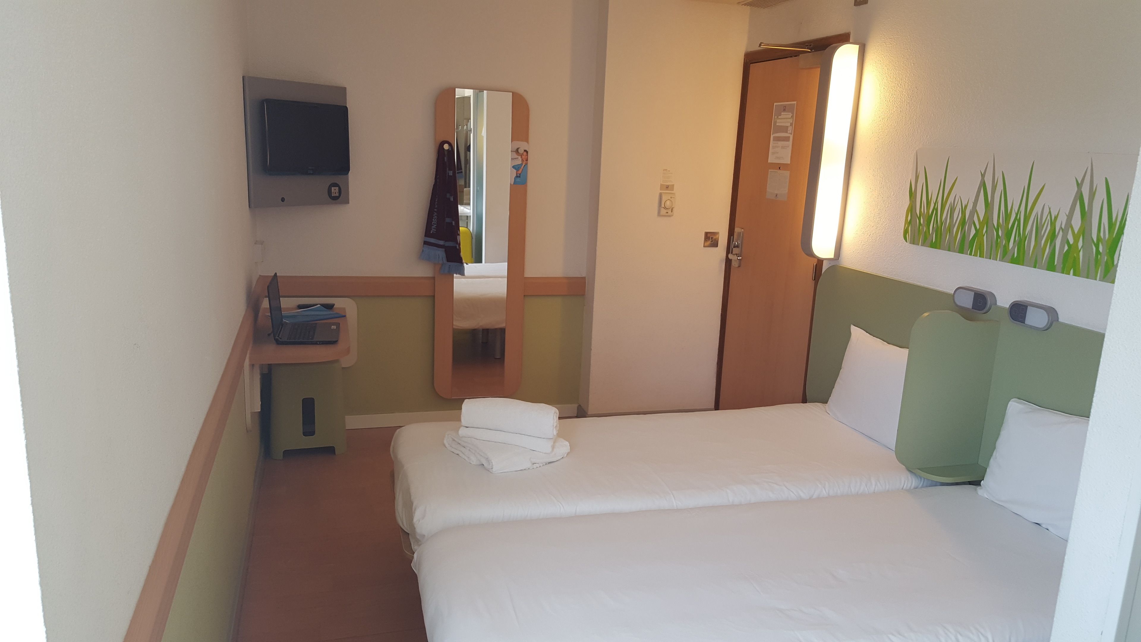 A hotel room with a bed, a desk with a laptop, a mirror, a door, and a wall-mounted TV.