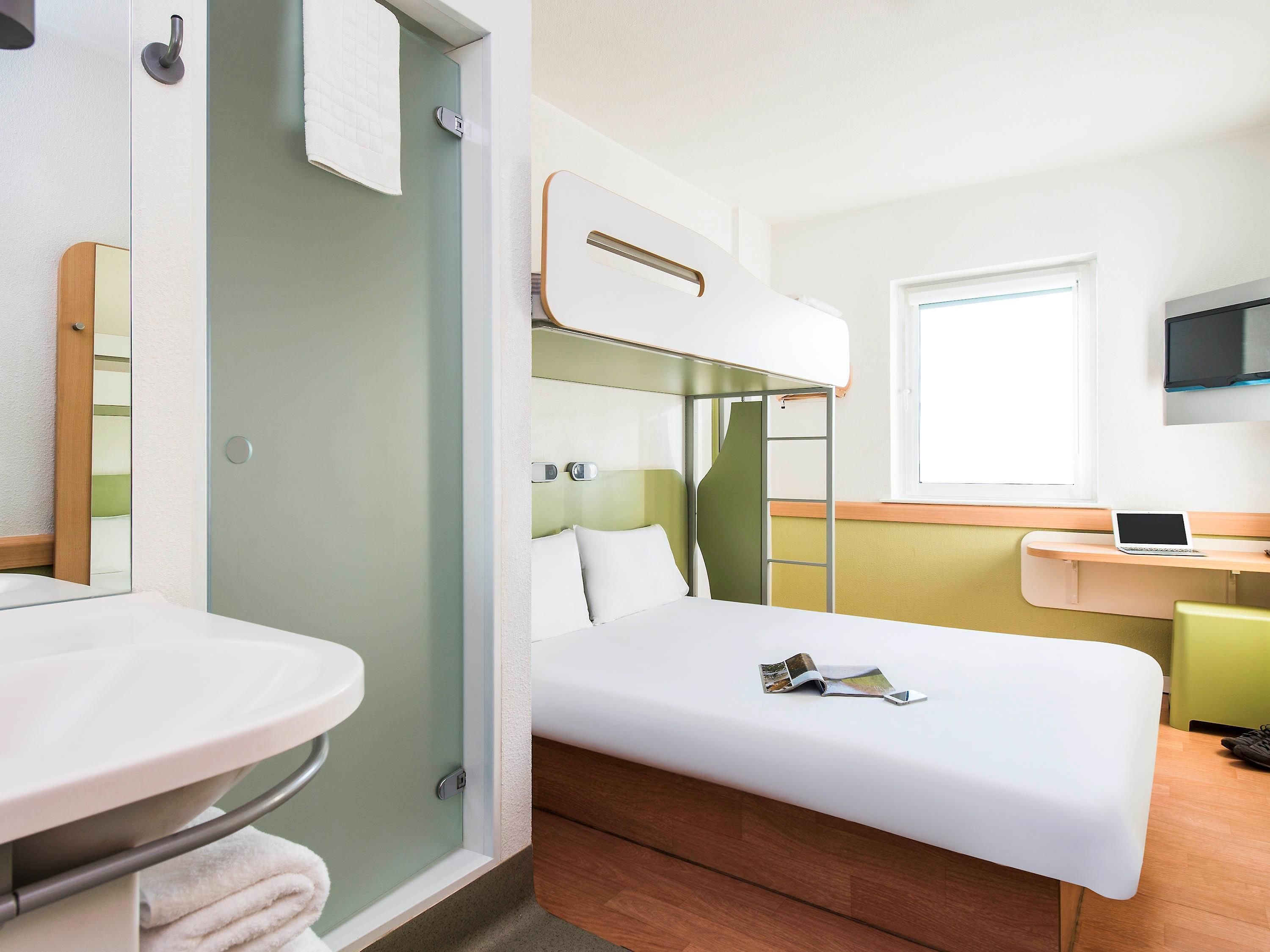 Ein kompaktes Hotelzimmer mit Etagenbett, einem kleinen Schreibtisch inklusive Laptop, einem Badezimmer mit Waschbecken und Spiegel sowie einem Fenster für natürliches Licht.