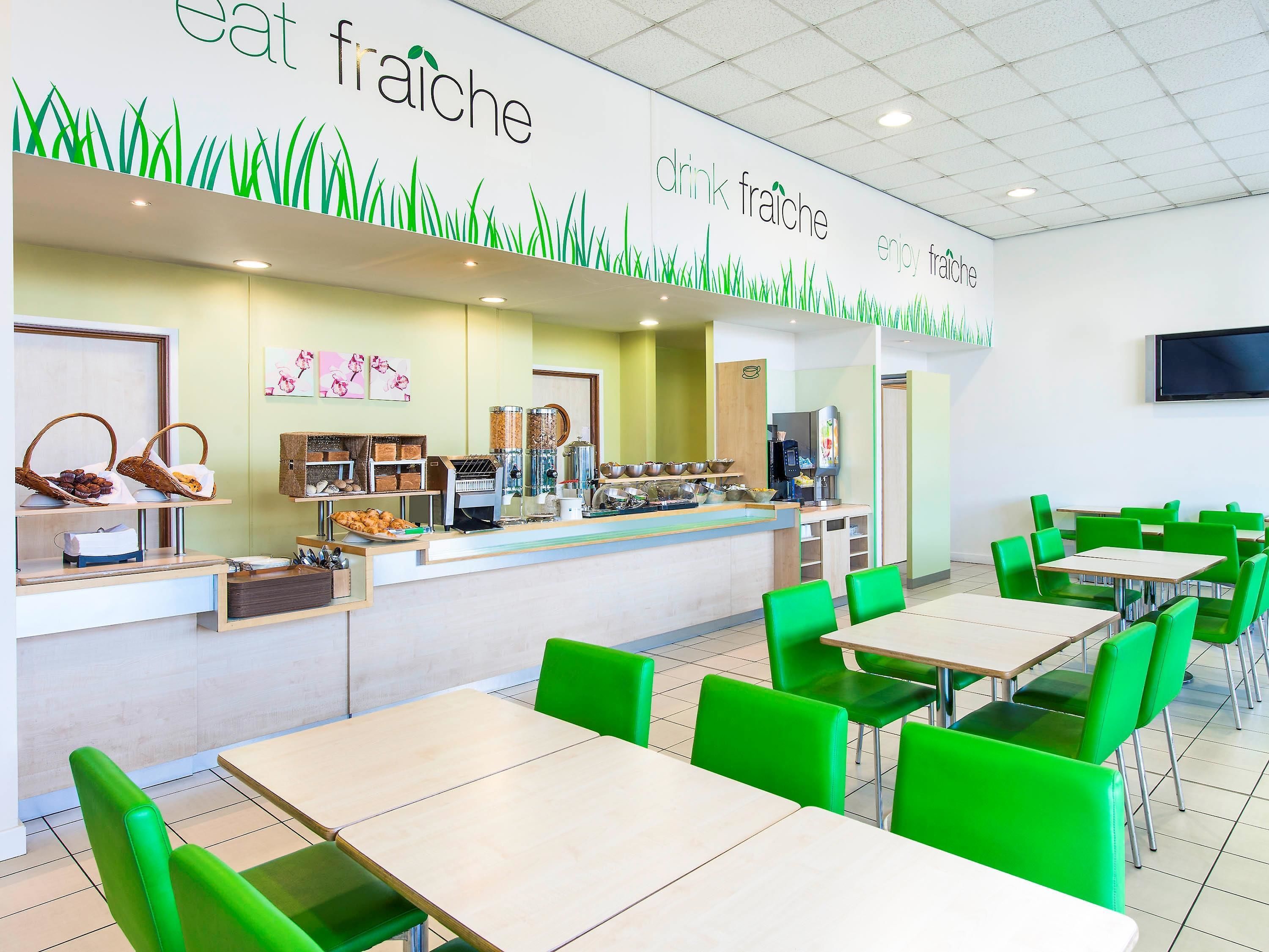 Una cafetería con un mostrador que exhibe alimentos y un área de estar con sillas y mesas verdes.