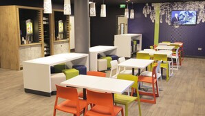 Restaurante moderno com mesas brancas e cadeiras coloridas, área de bar e uma TV exibindo um noticiário.