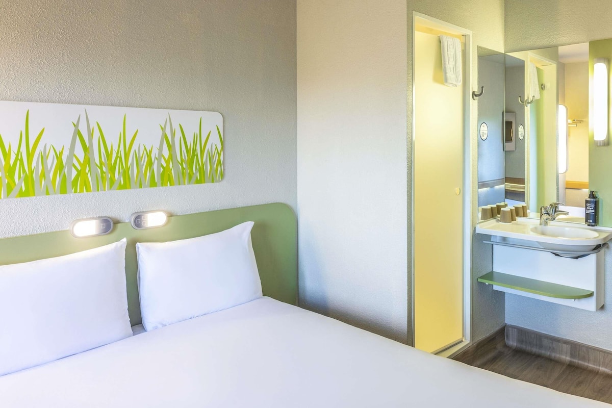 Una habitación de hotel con cama, un cuadro de césped colgado en la pared, un baño con lavamanos y espejo, y un estante verde.
