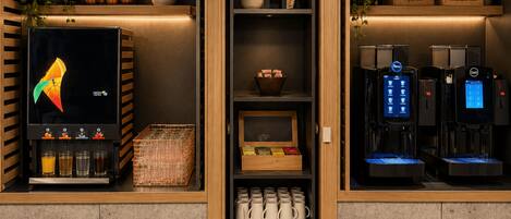 Interior de una cafetería con una máquina de café, una exposición de frutas y una estantería con tazas.