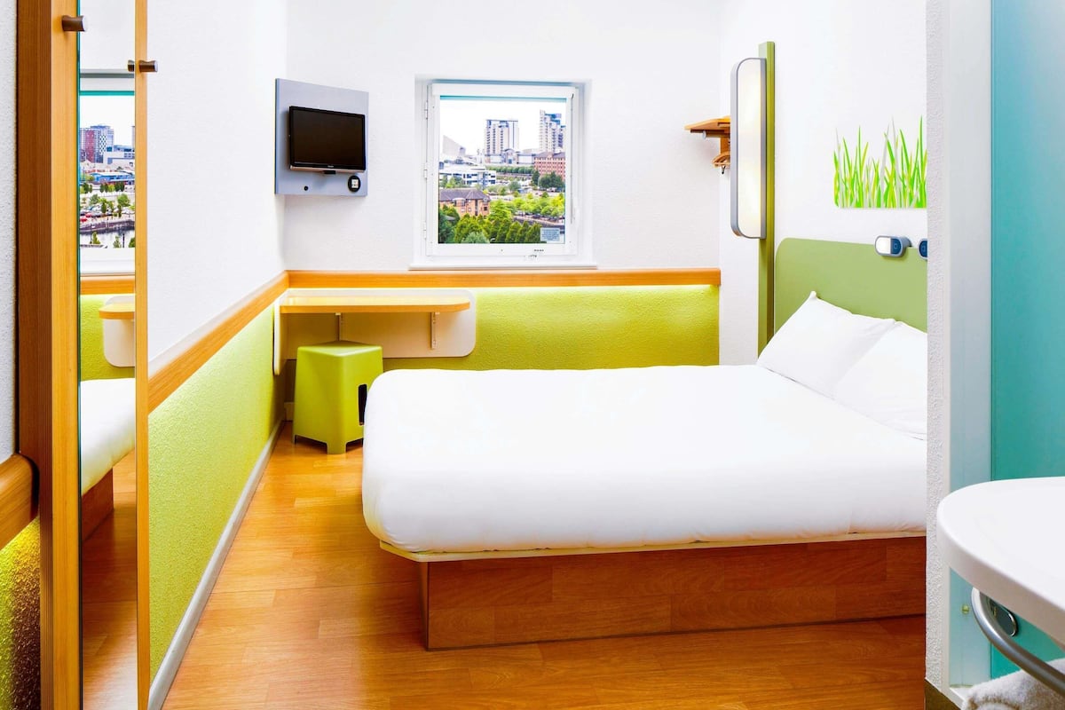 Una camera d'albergo con un letto, una scrivania, una televisione, una finestra vista città e un bagno.