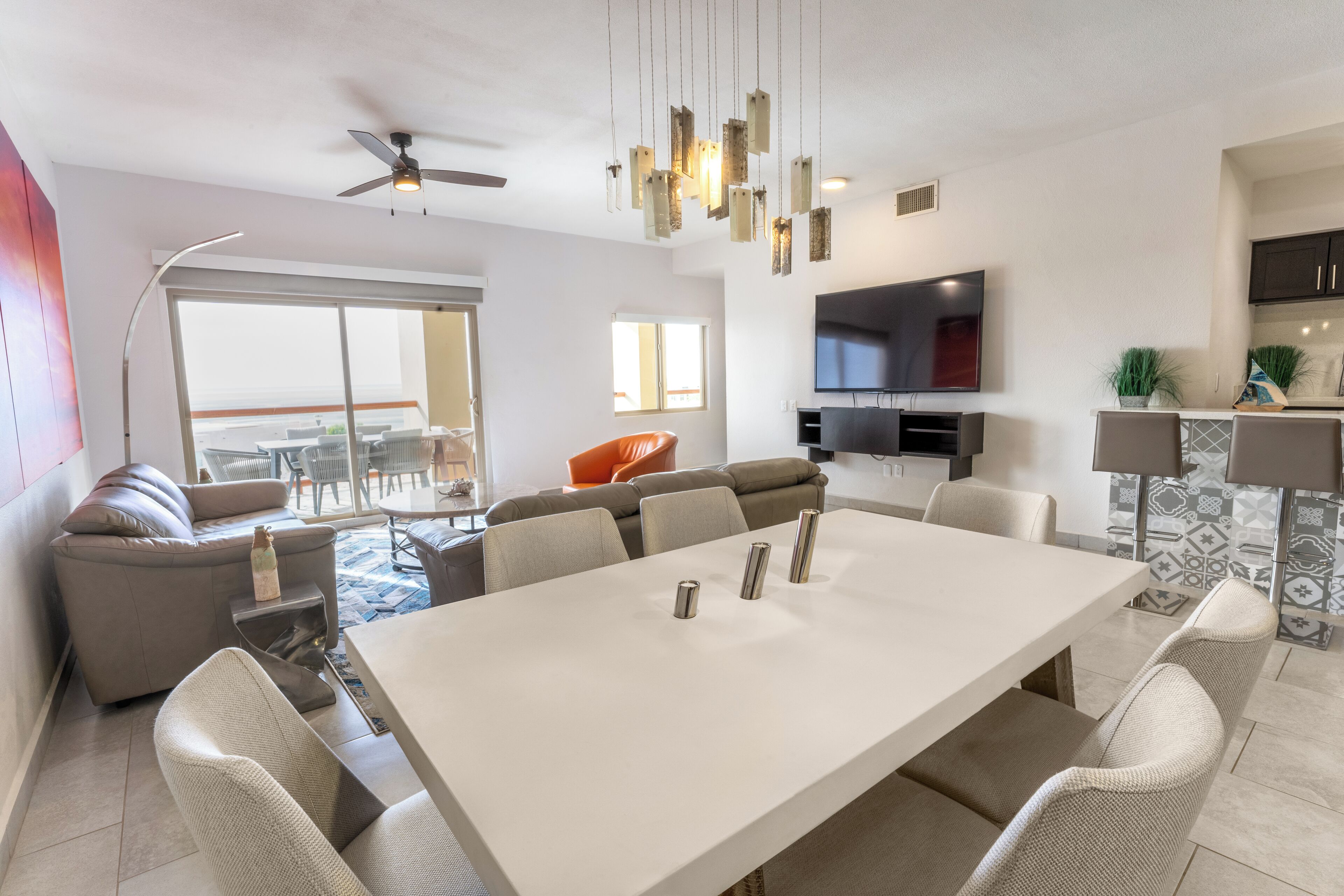 Un salon moderne avec une grande table à manger blanche, des canapés gris et un téléviseur à écran plat fixé au mur.