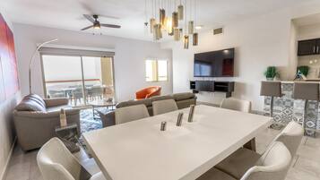 A modern living room with a large white dining table, grey sofas, and a flat-screen TV mounted on the wall.