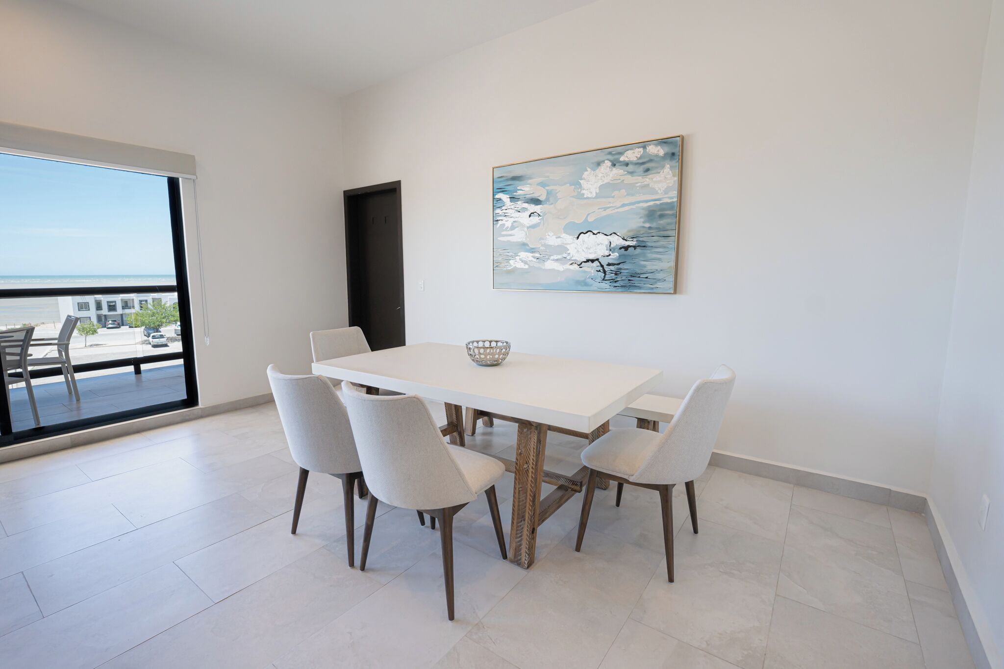 A dining area with a white table and four chairs, a bowl on the table, and a painting on the wall.