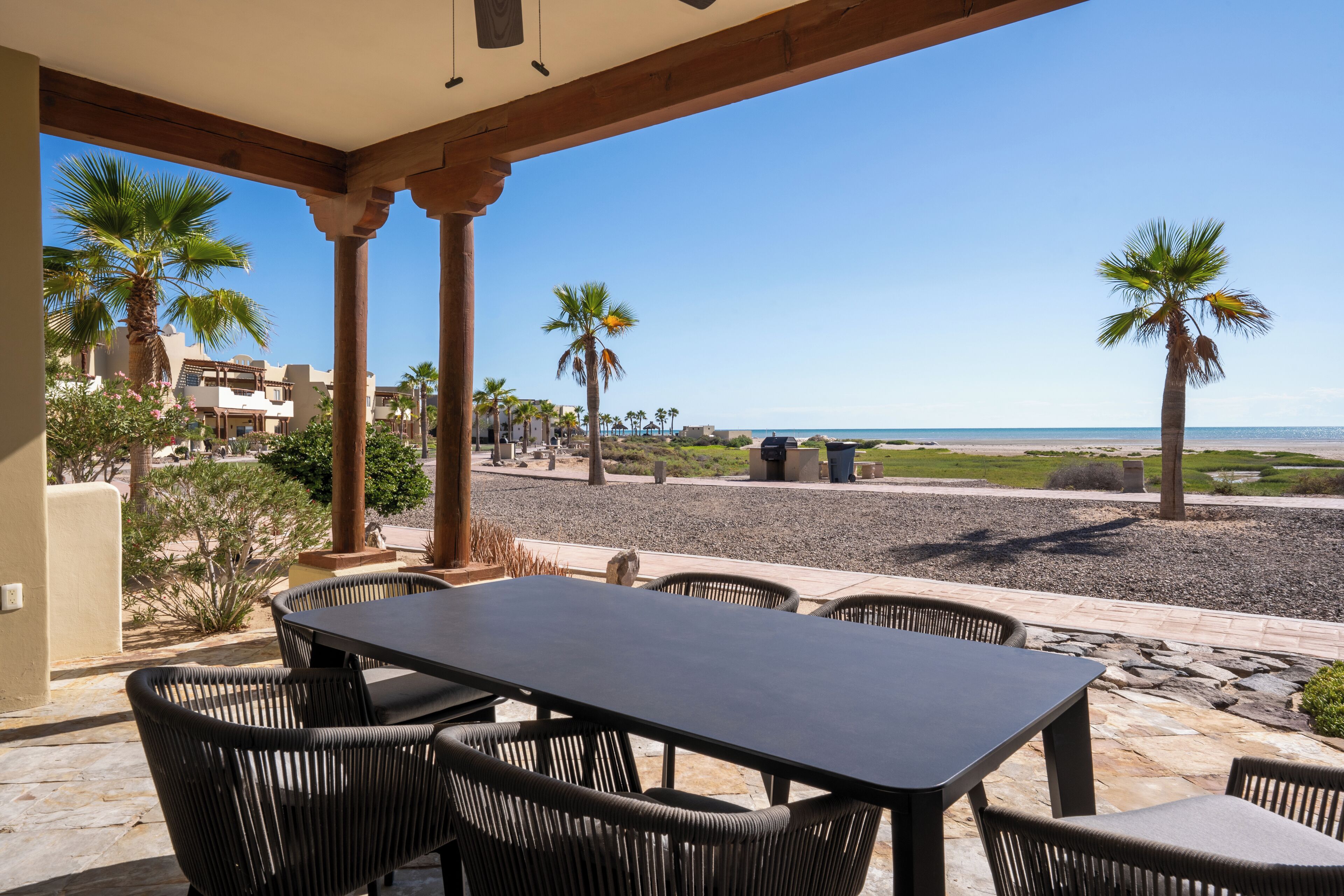 An outdoor patio with a table and chairs, overlooking a beach with palm trees and a clear sky.