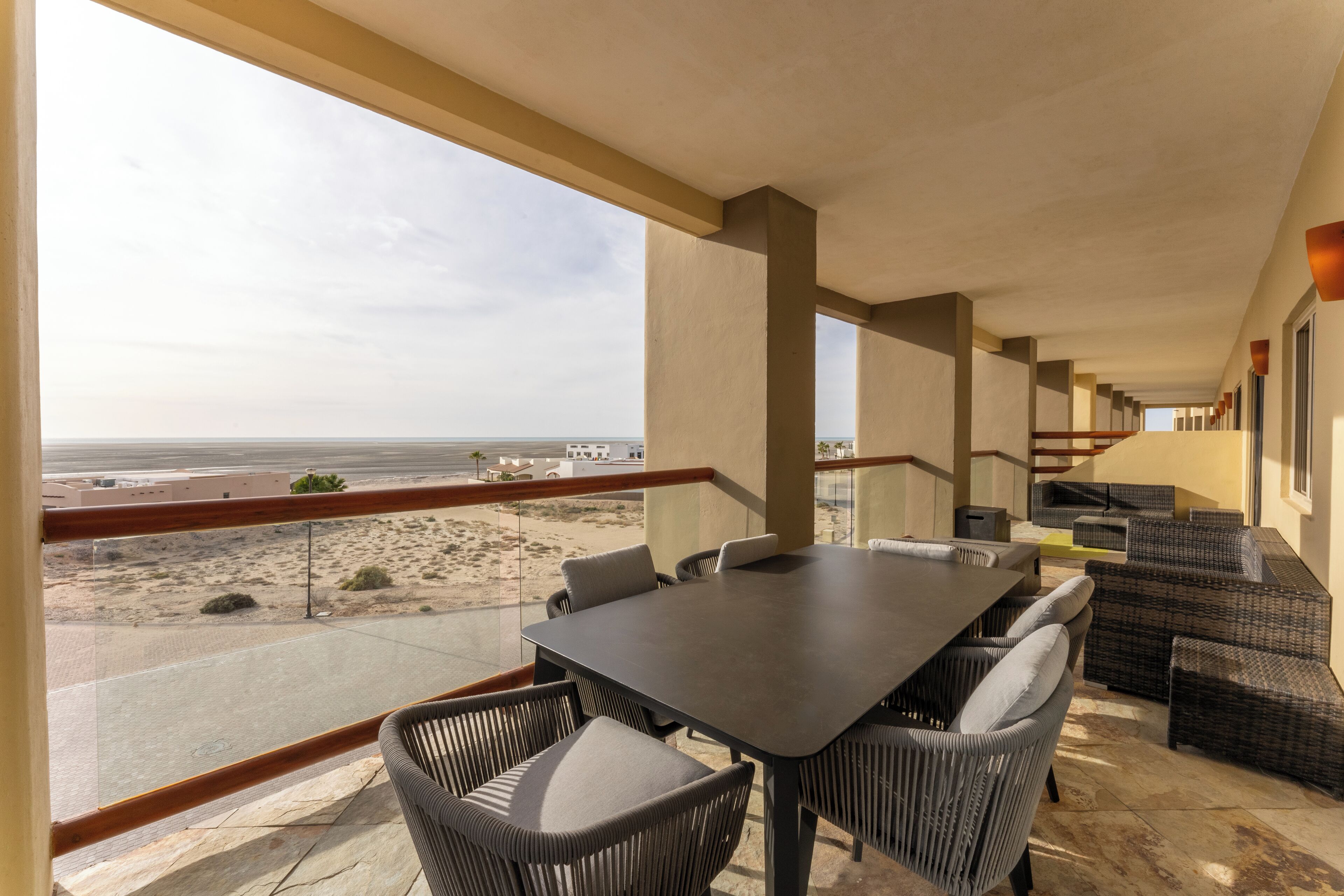 Un balcon avec une table et des chaises, donnant sur un paysage désertique avec une route et des bâtiments visibles au loin.