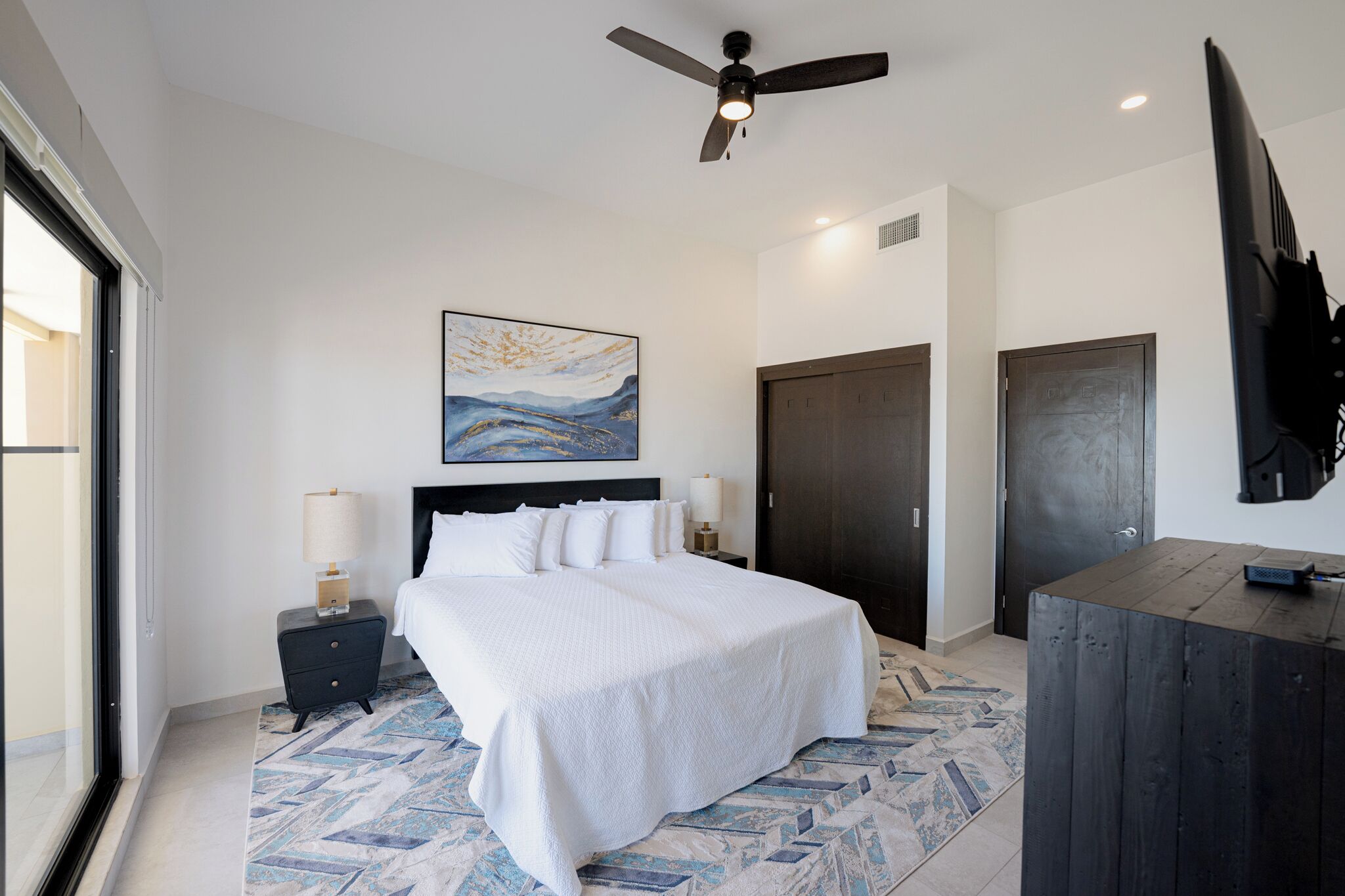 A modern hotel room with a large bed, a ceiling fan, a painting, a dark door, and a flat-screen TV.