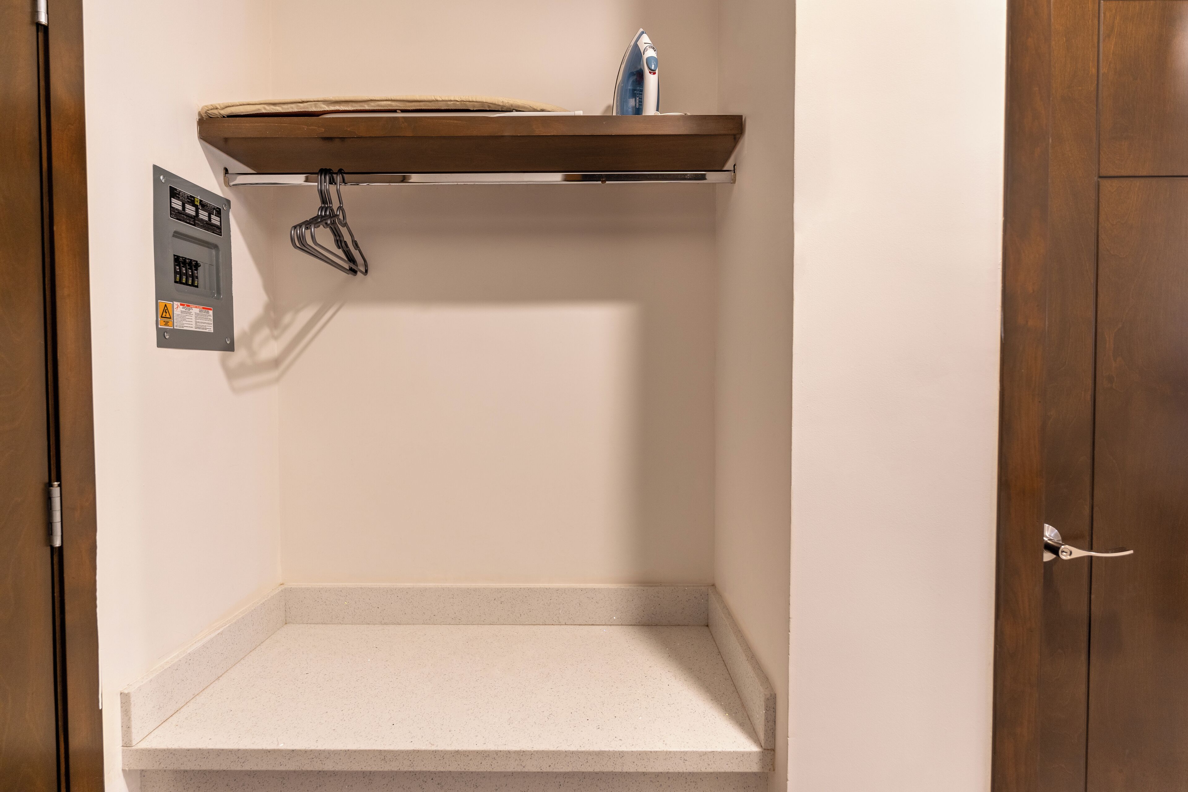 A bathroom with a wooden shelf above a countertop, a towel hanging on a hook, and a power outlet with a warning label.