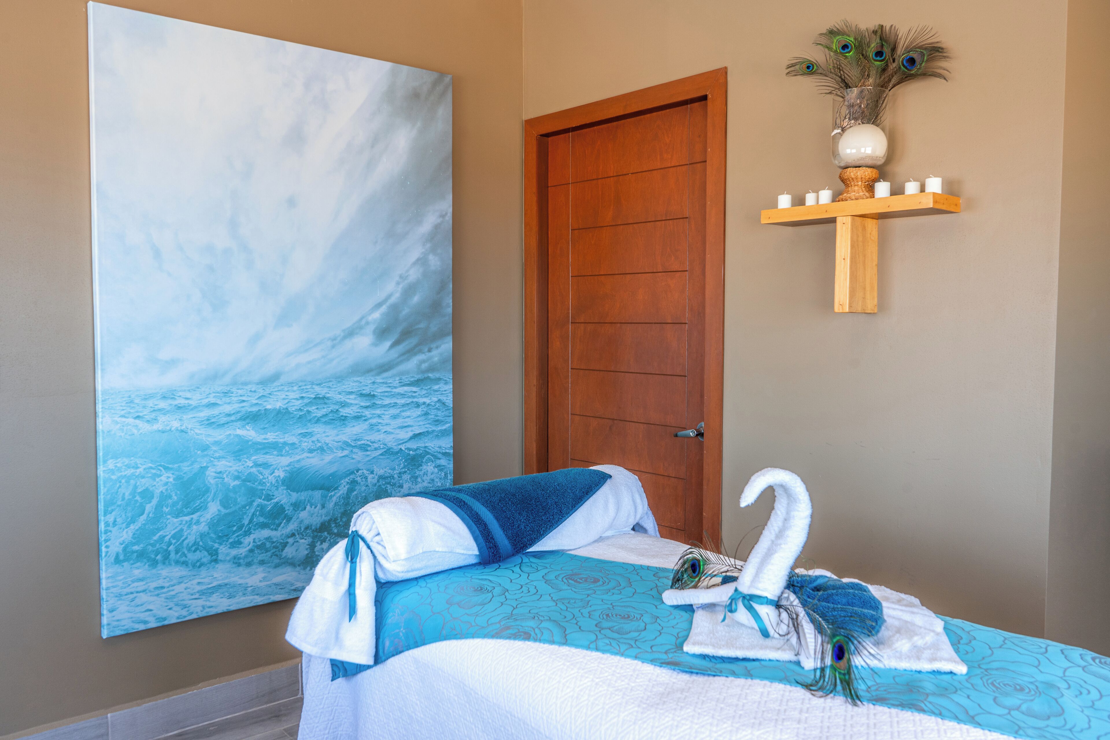 A hotel room with a large ocean-themed painting, a wooden door, and a shelf with a vase and candles.