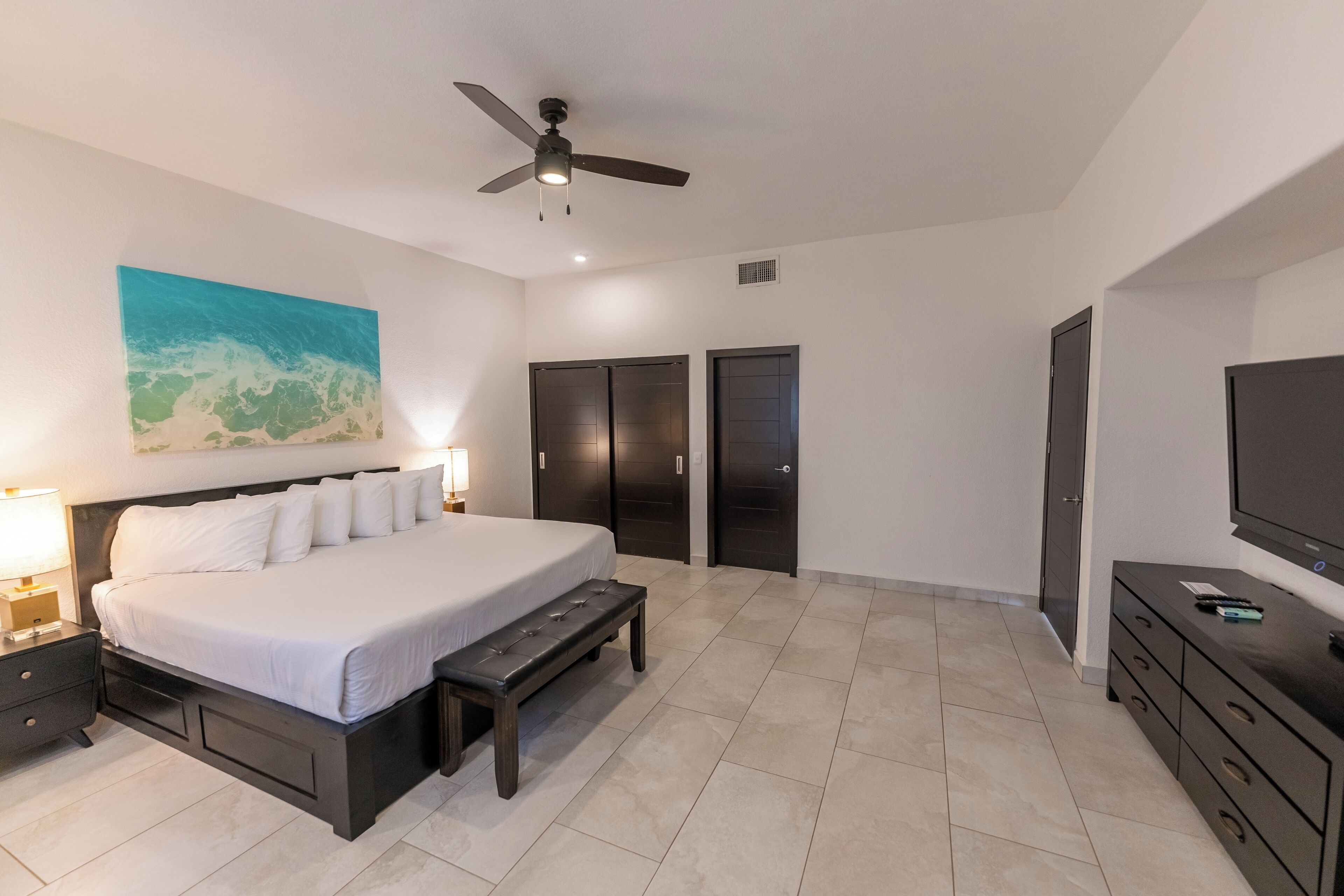 A bedroom with a bed, bedside tables, a bench, a ceiling fan, a TV, and a painting on the wall.