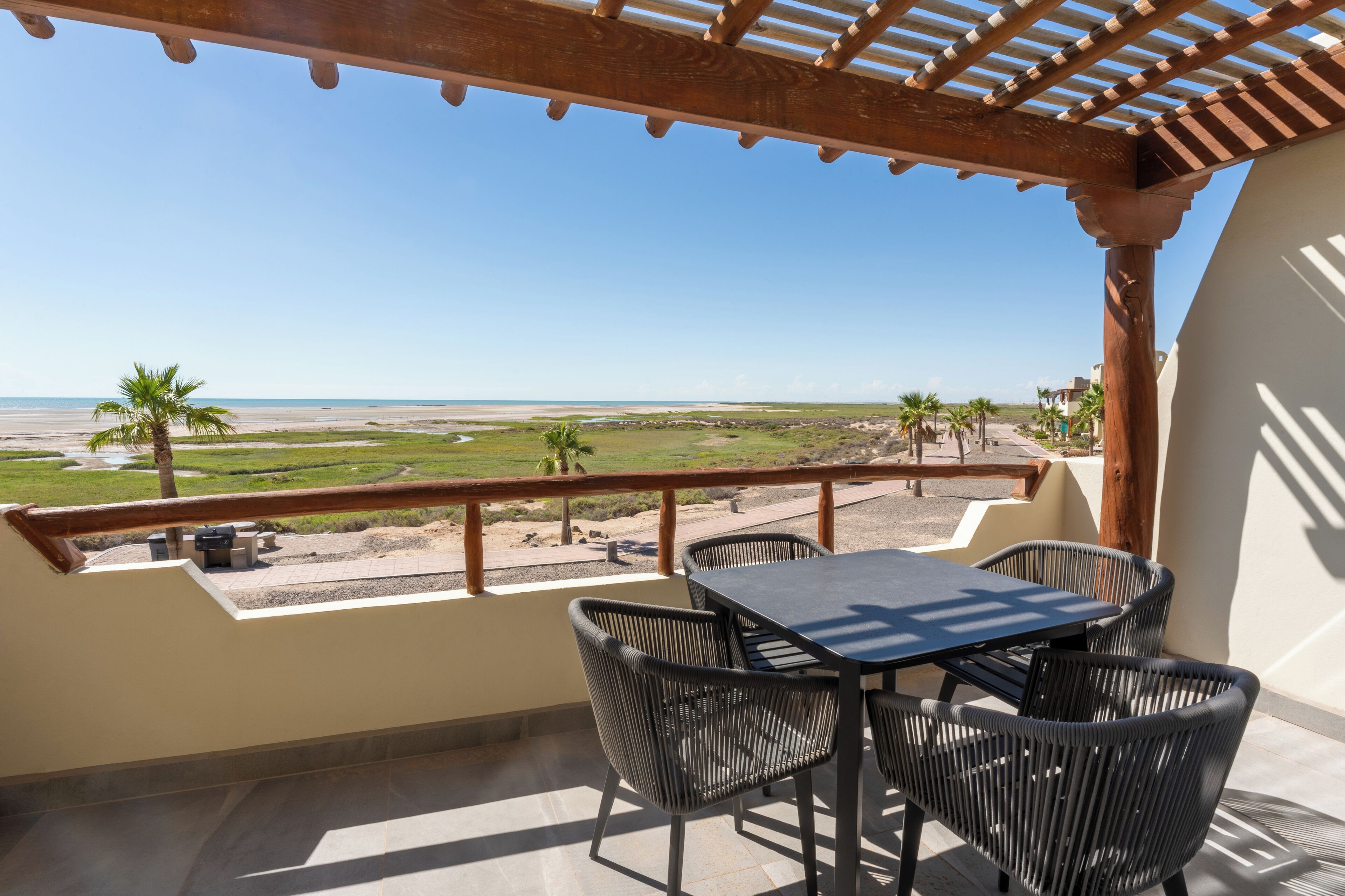 Balcón con mesa y sillas, con vista a una playa y palmeras.