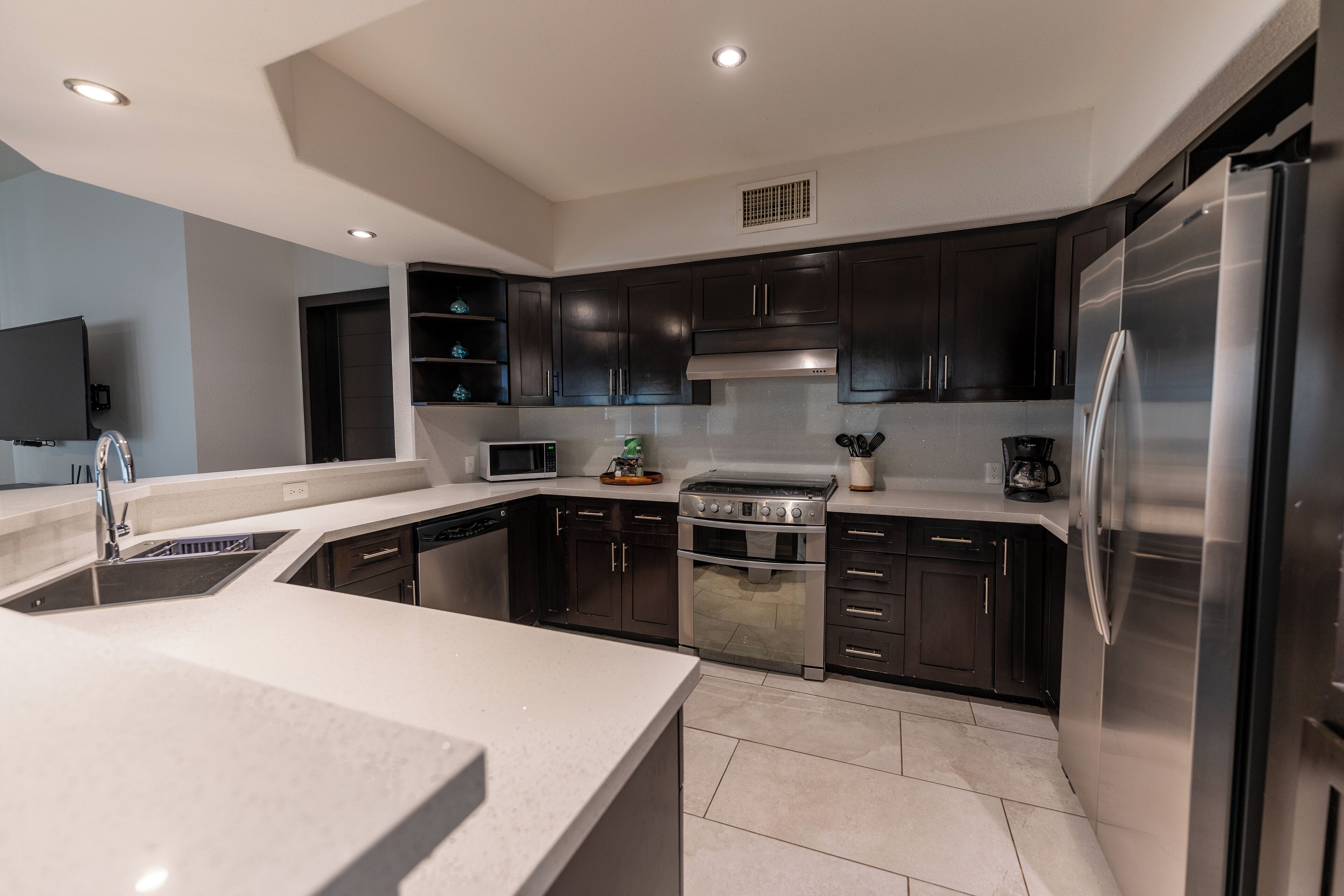 A modern kitchen with dark cabinets, stainless steel appliances, and a flat-screen TV mounted on the wall.