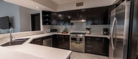 A modern kitchen with dark cabinets, stainless steel appliances, and a flat-screen TV mounted on the wall.