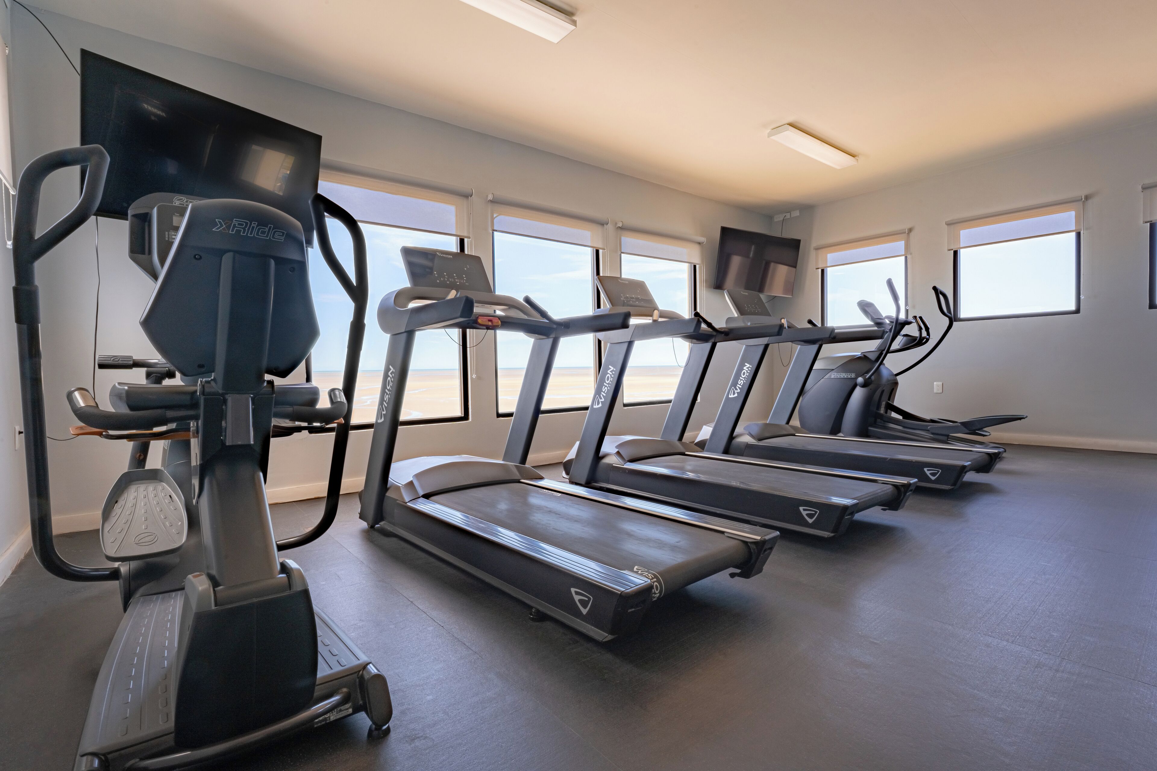Ein Fitnessstudio mit Laufbändern und Crosstrainern, großen Fenstern und einem ebenen Boden.