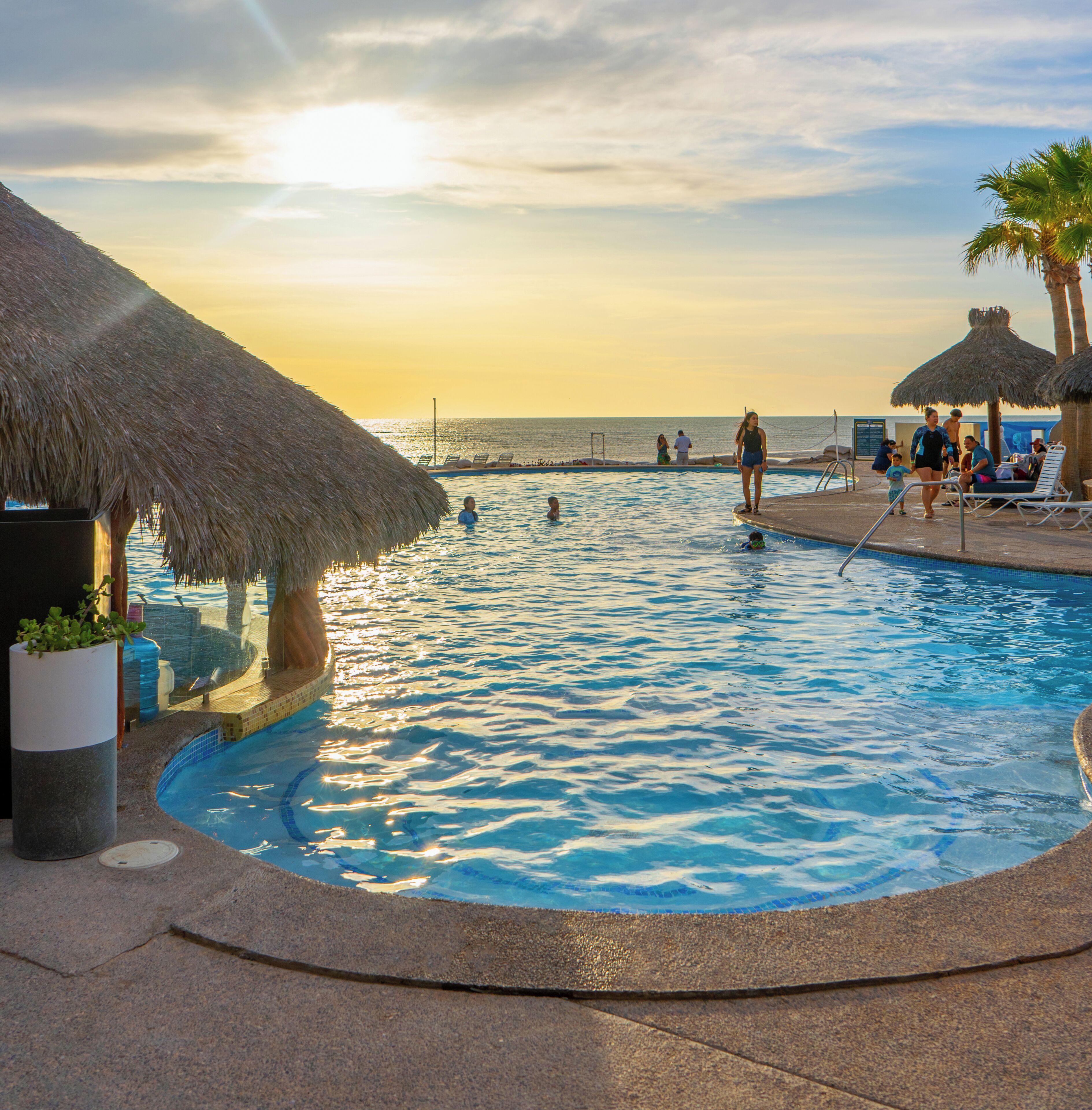 A swimming pool with a curved edge, a thatched roof structure, and a view of the sea in the background.
