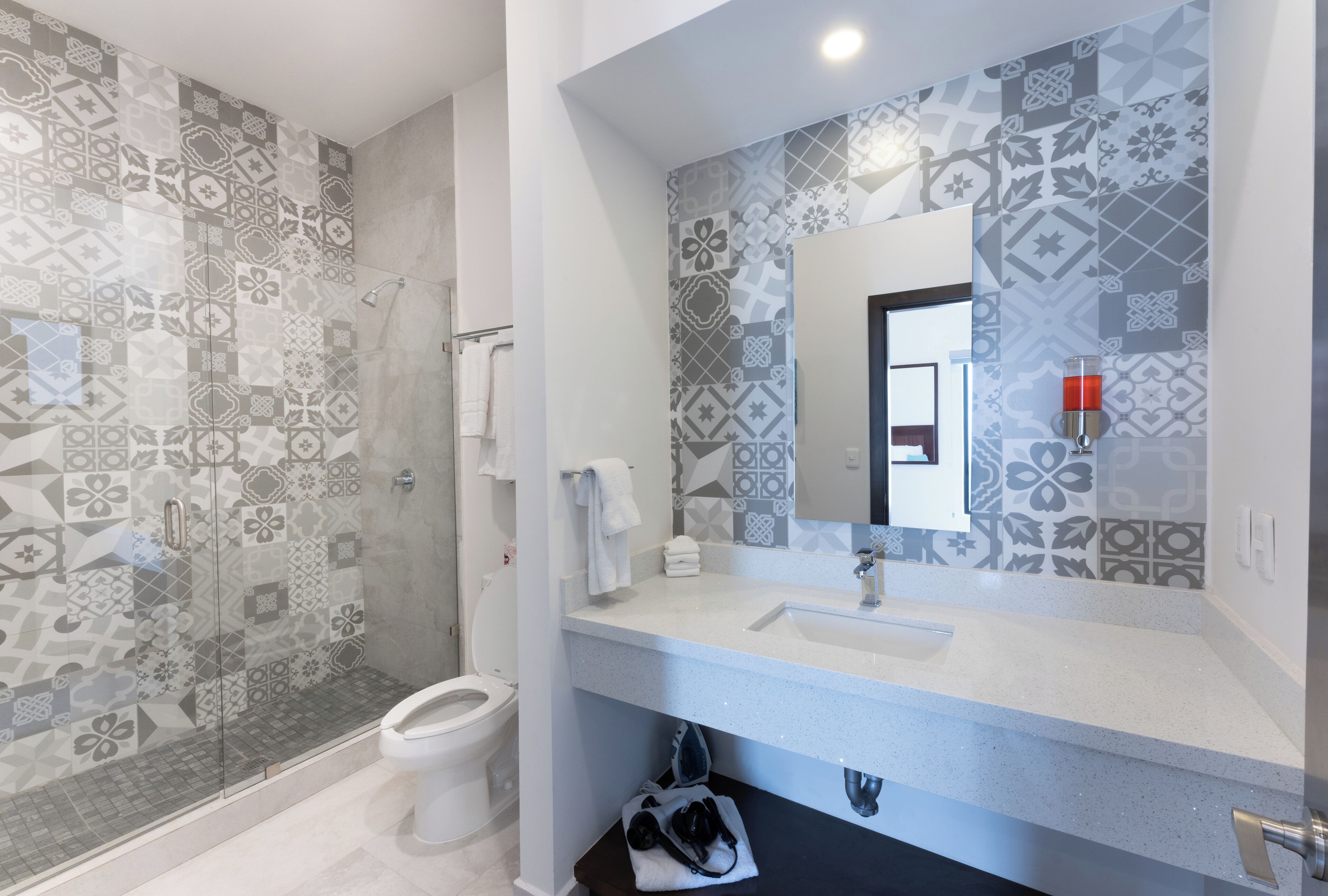 A bathroom with patterned tiles, a glass shower enclosure, a white sink, and a mirror with a red light.
