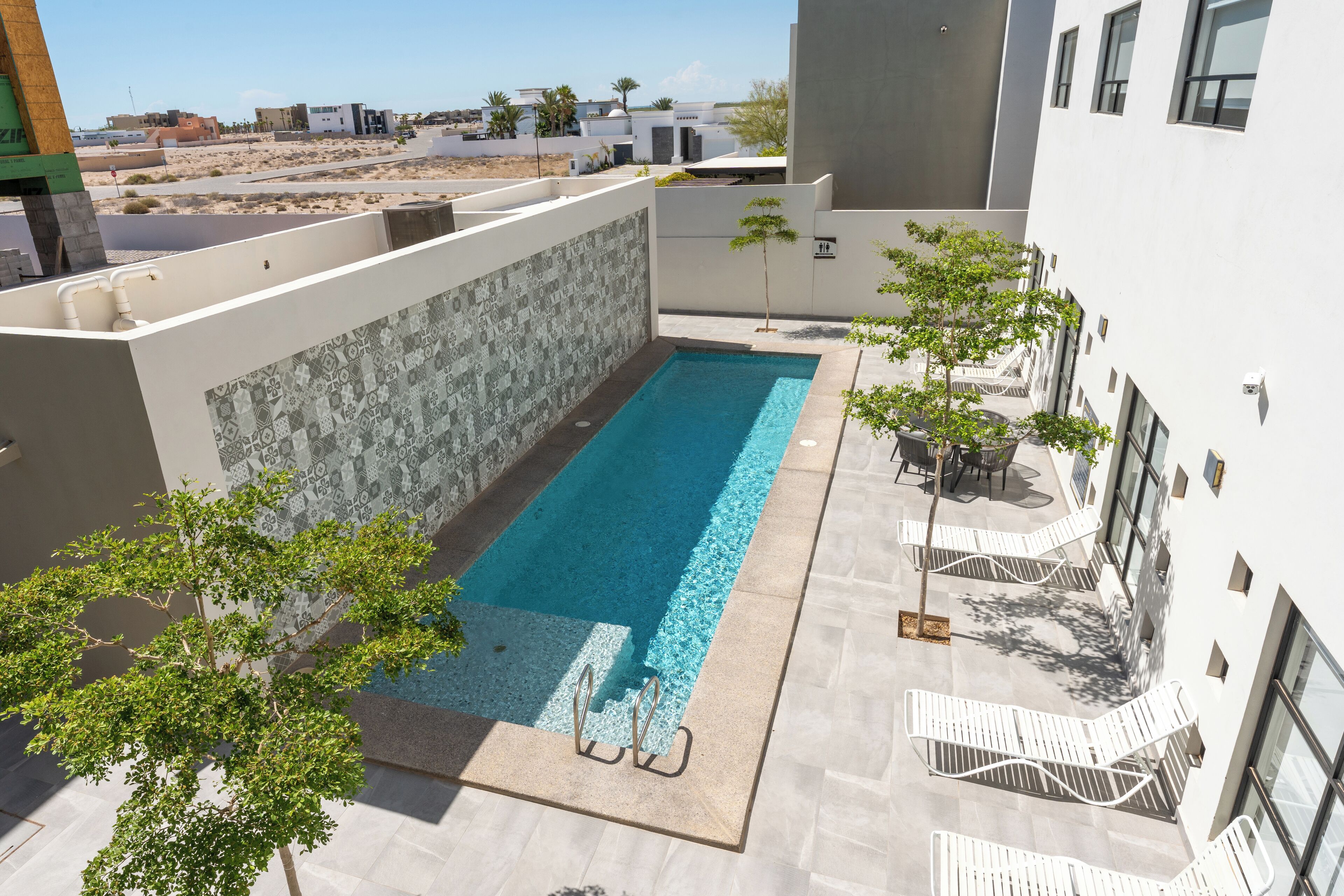 A rooftop pool with a tiled deck, surrounded by white walls and a few trees.