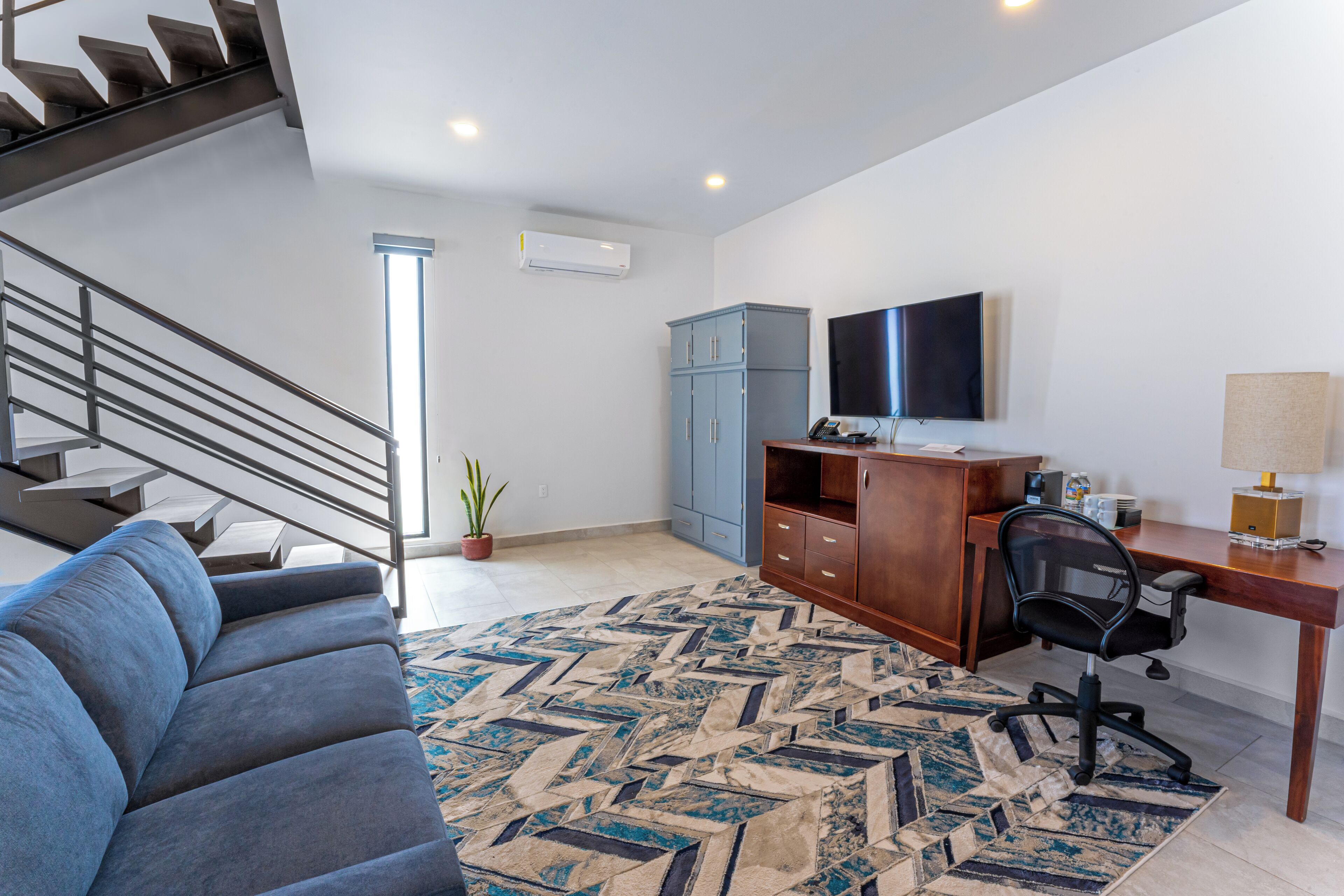 Un salon moderne avec un escalier, un canapé bleu, un bureau en bois, un ordinateur et un téléviseur.