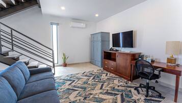 A modern living room with a staircase, a blue sofa, a wooden desk, a computer, and a TV.
