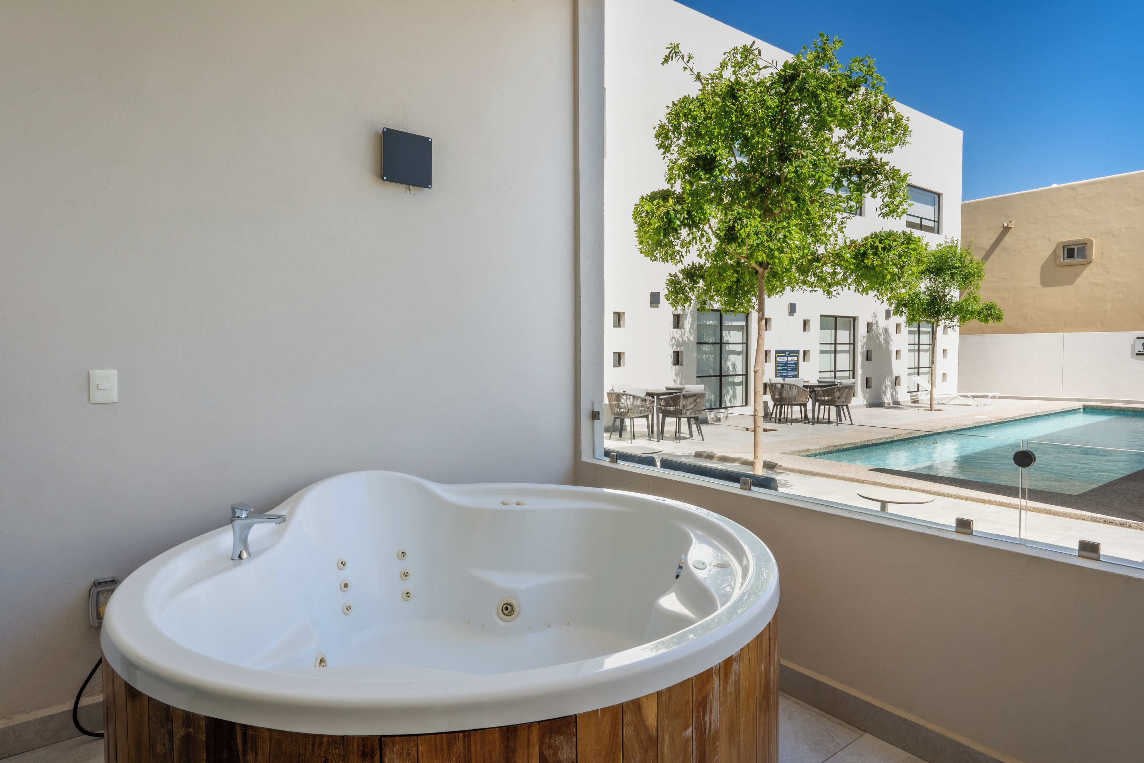 A modern bathroom with a freestanding bathtub, a window overlooking a pool area, and a view of a building with outdoor seating.