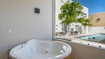 A modern bathroom with a freestanding bathtub, a window overlooking a pool area, and a view of a building with outdoor seating.