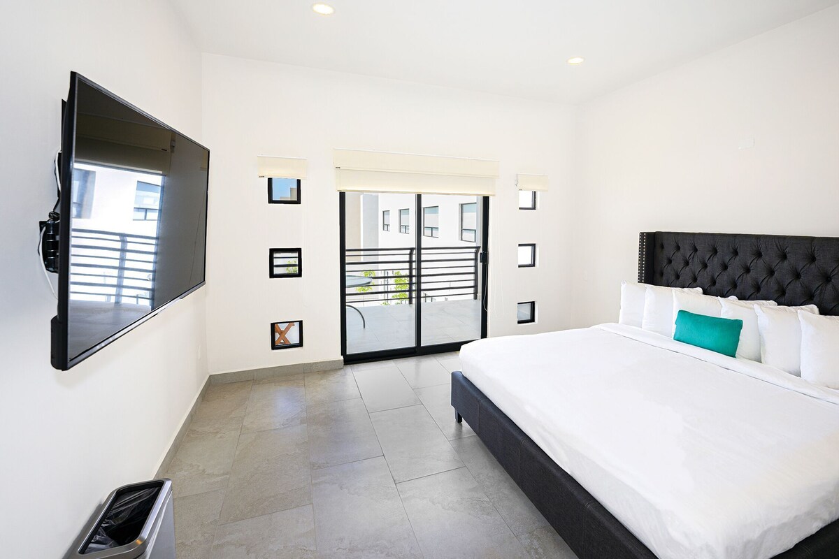 Ein modernes Hotelzimmer mit einem großen Bett, einem flachbildfernseher an der Wand, einem Balkon mit Aussicht und einer Schiebetür zu einem Außenbereich.