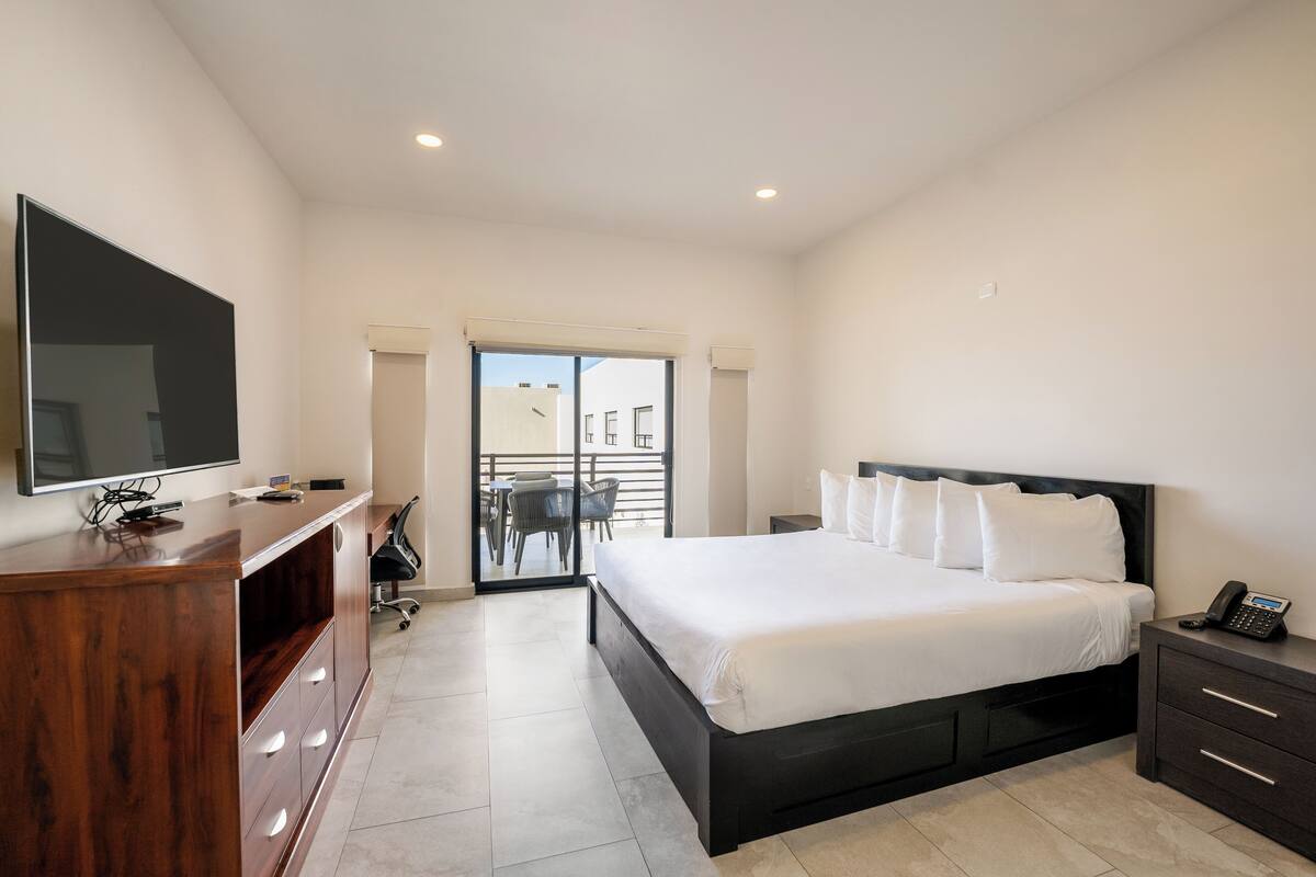 Een moderne hotelkamer met een groot bed, een houten bureau, een flatscreen televisie en een balkon met zitgelegenheid buiten.