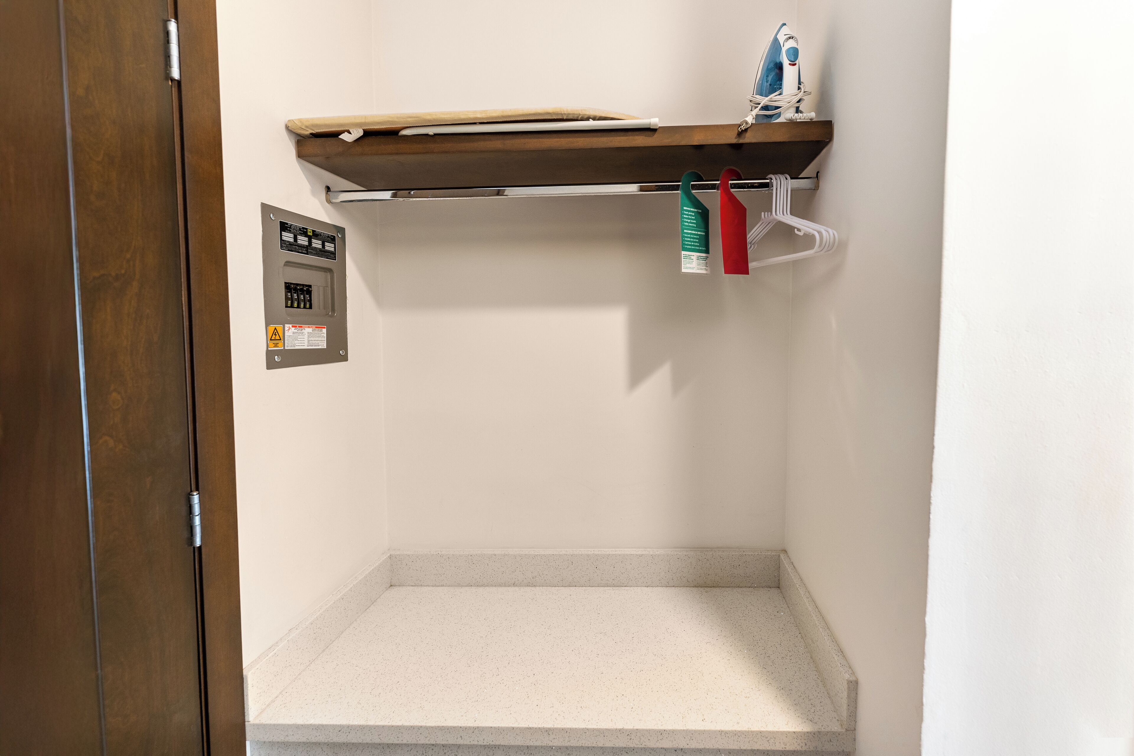 A hotel room with a wooden door, a control panel, a shelf with an iron, and a hanging rack with clothes.