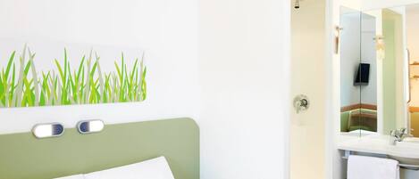 Hotelkamer met een bed, een wandspiegel, een badkamer met een wastafel en handdoekhouder, en een groene muur met grasdecoratie.