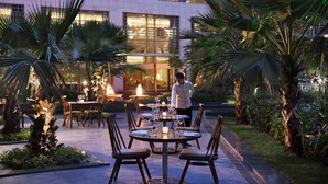An outdoor dining area with tables and chairs, surrounded by palm trees and a building in the background.
