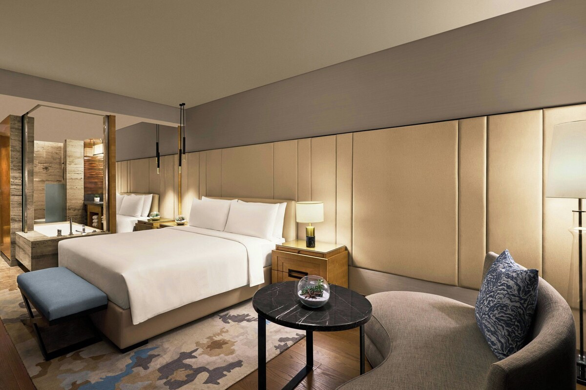 Una habitación de hotel con cama, zona de estar, mesita de noche, lámpara y espejo.