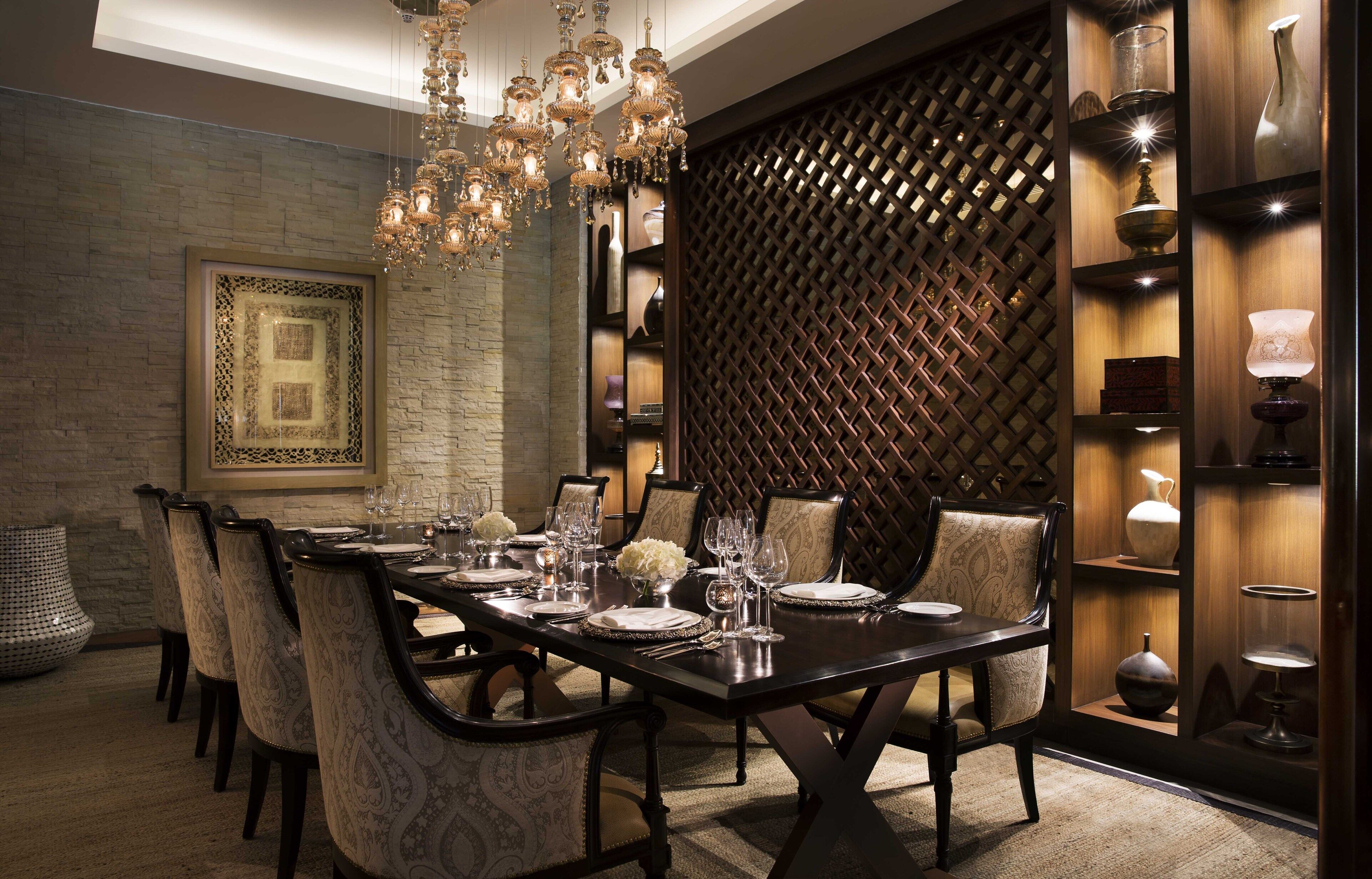 Un comedor con una larga mesa dispuesta para seis personas, rodeada de sillas con diseños intrincados. Sobre ella, un candelabro grande y ornamentado, y a la derecha, un panel decorativo con diseño de celosía.