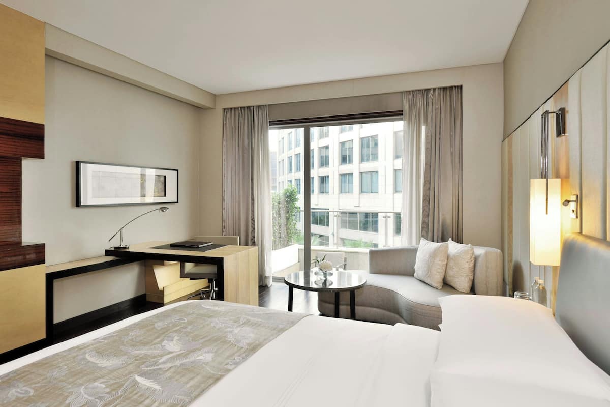 A hotel room with a large bed, a desk with a chair, a sofa, a small table with a vase, and a window with curtains.