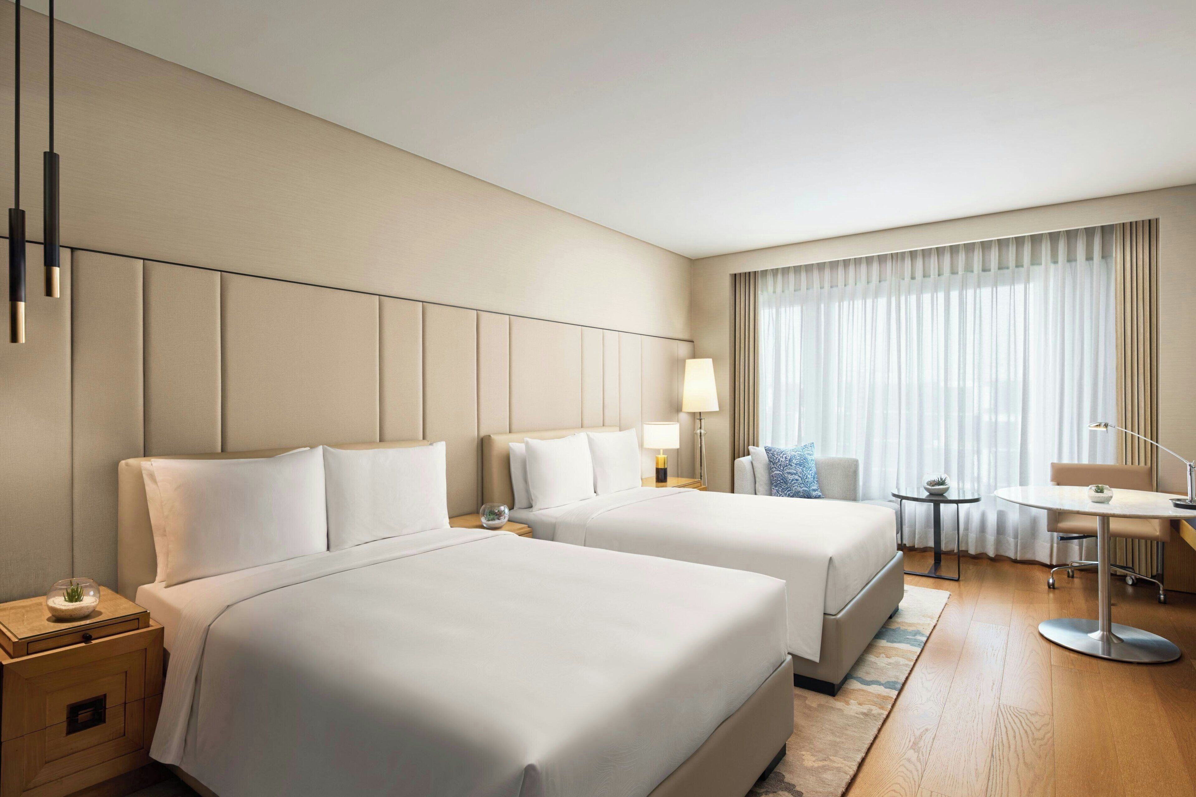A hotel room with two beds, a desk, a chair, a lamp, and a window with curtains.