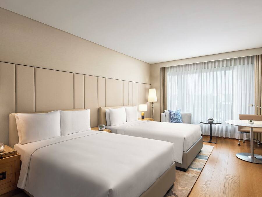 A hotel room with two beds, a desk, a chair, a lamp, and a window with curtains.