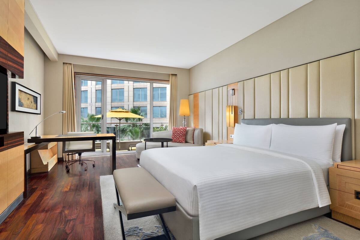 A hotel room with a large bed, a desk, a chair, a bench, a nightstand, and a window with a view of buildings and greenery.