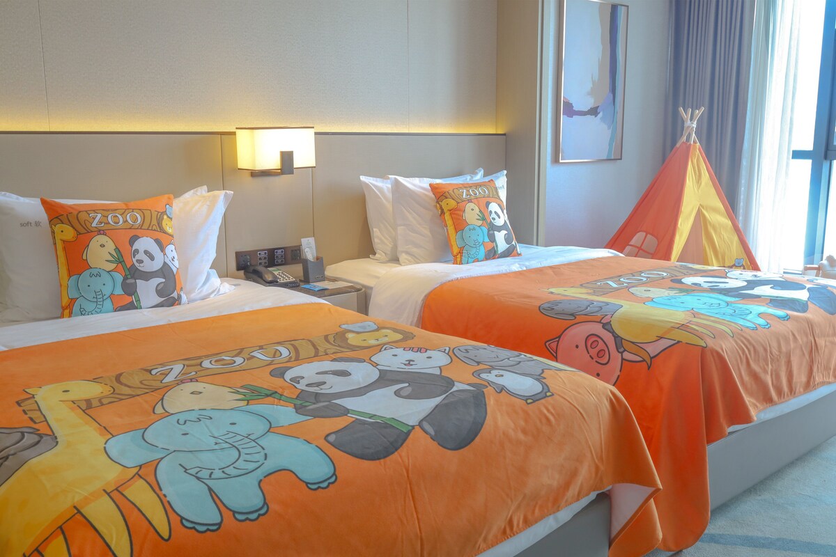 A hotel room with two beds, each with an orange bedspread featuring cartoon animals and the word "ZOO". There is a nightstand with a lamp and a phone, and a window with curtains.