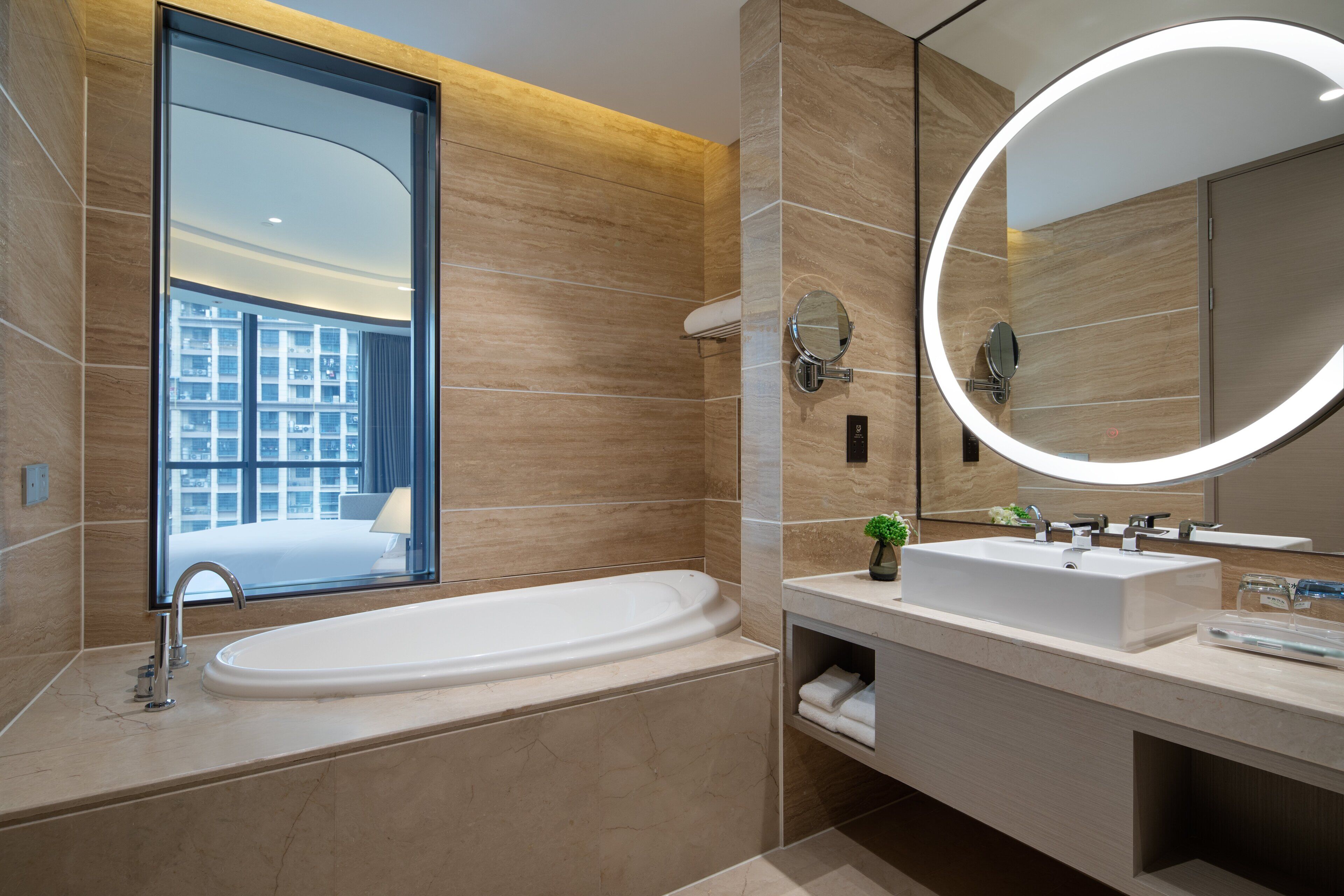 Une salle de bain moderne avec une grande baignoire, un grand miroir rond doté d’un éclairage intégré et une fenêtre donnant sur un immeuble.