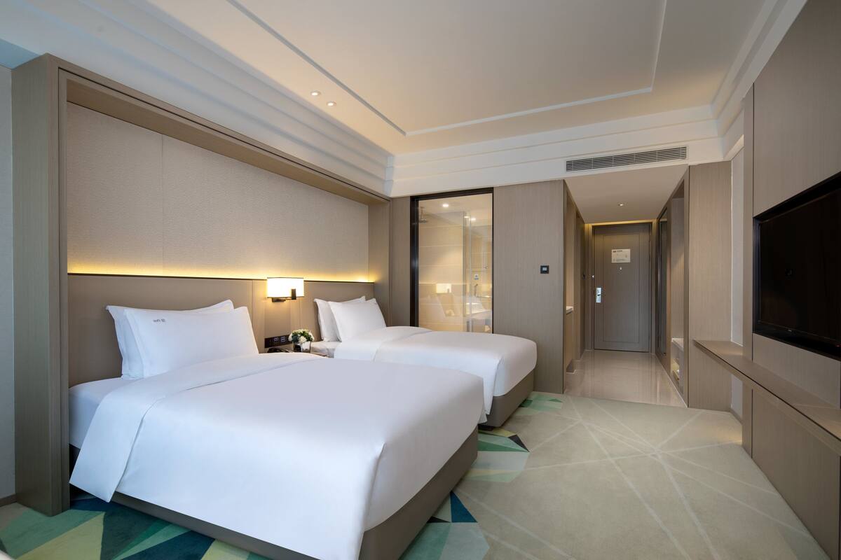 Ein Hotelzimmer mit zwei Betten, einem Fernseher, einem Nachttisch und einem durch eine Glastür sichtbaren Badezimmer.
