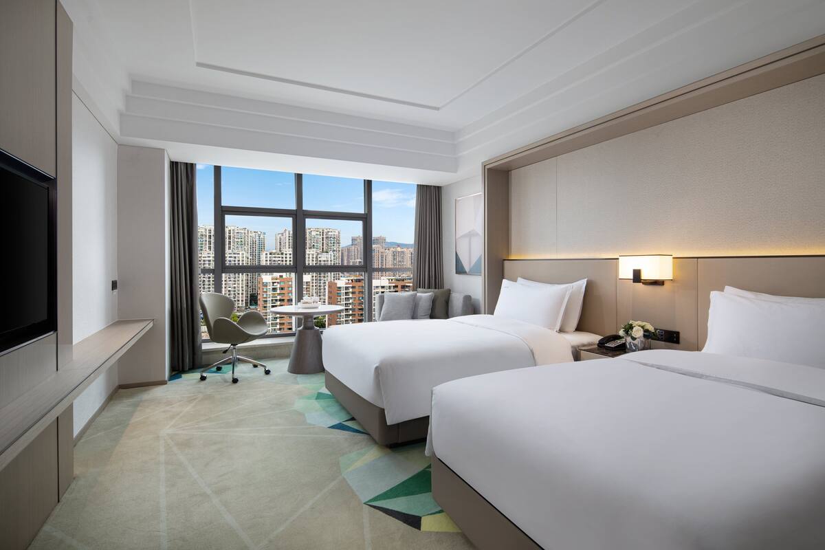 Ein Hotelzimmer mit zwei Betten, einem Fernseher, einem Schreibtisch und Blick auf die Stadt.