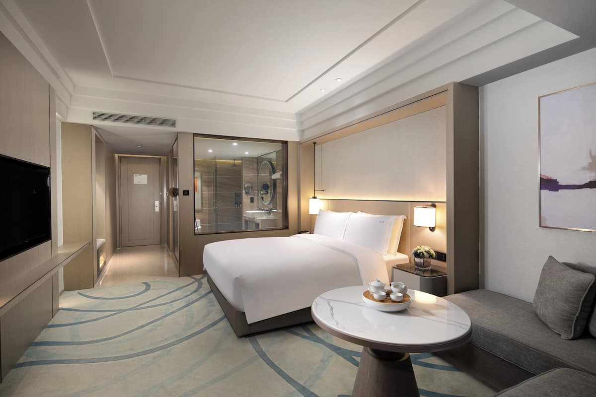 Ein modernes Hotelzimmer mit einem großen Bett, einem Flachbildfernseher, einem Nachttisch mit Lampe und einem kleinen runden Tisch mit dekorativer Schale.