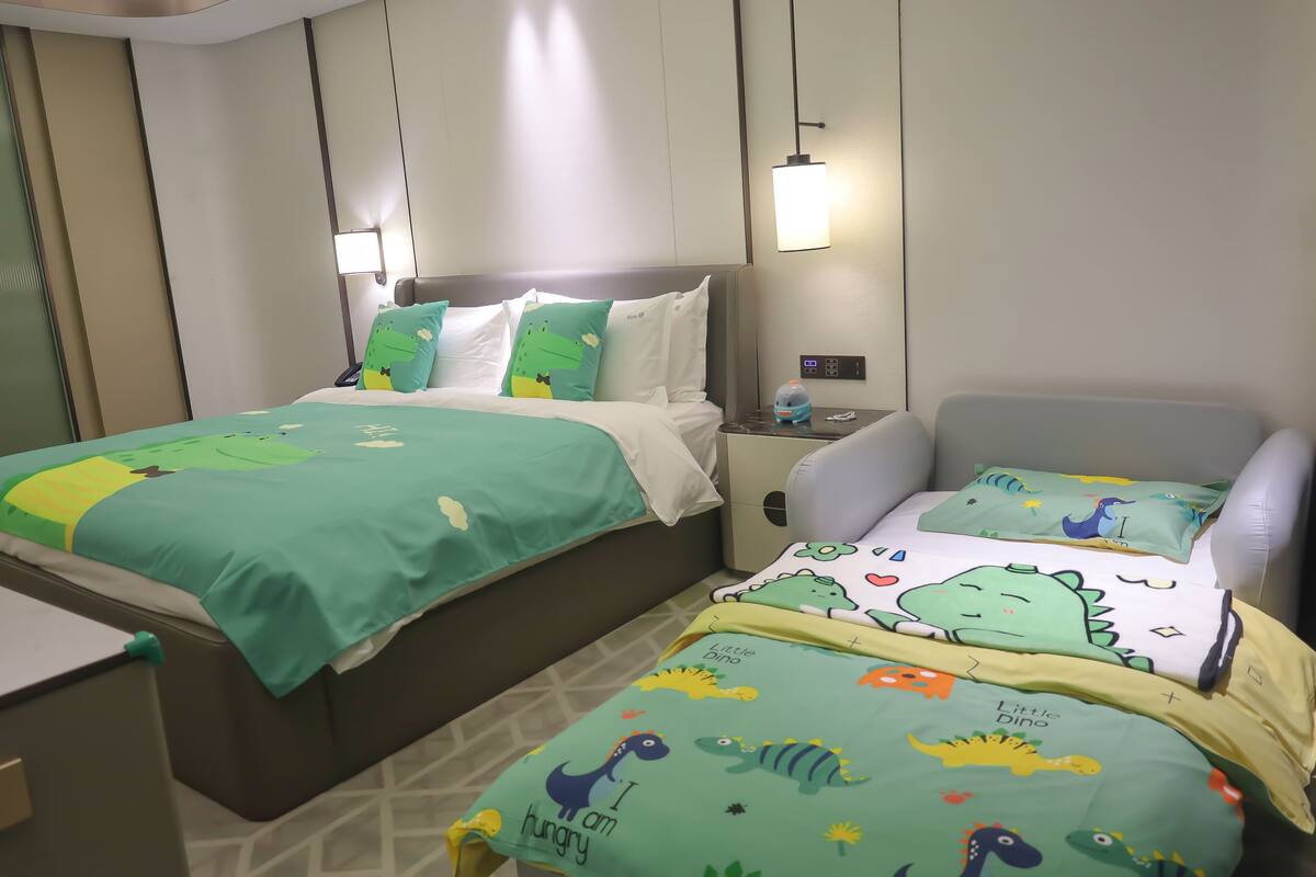 Ein Hotelzimmer mit zwei Betten, beide mit Bettwäsche im Dinosaurier-Design. Es gibt Nachttische mit Lampen und einen Beistelltisch mit einem Telefon.